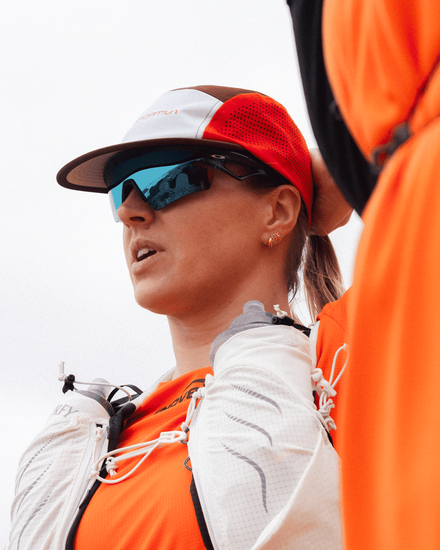 A person wearing a cap and mirrored sunglasses, dressed in orange mountain running gear, outdoors against a cloudy sky.