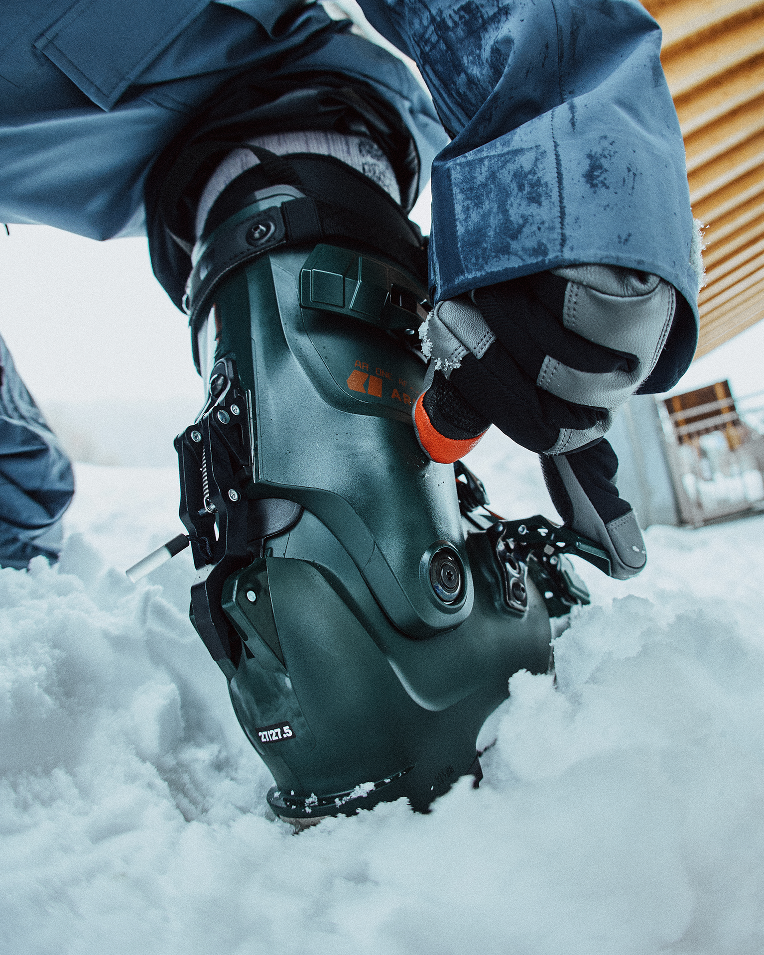 Person adjusting bindings on a green ski boot in the snow, winter gear visible.