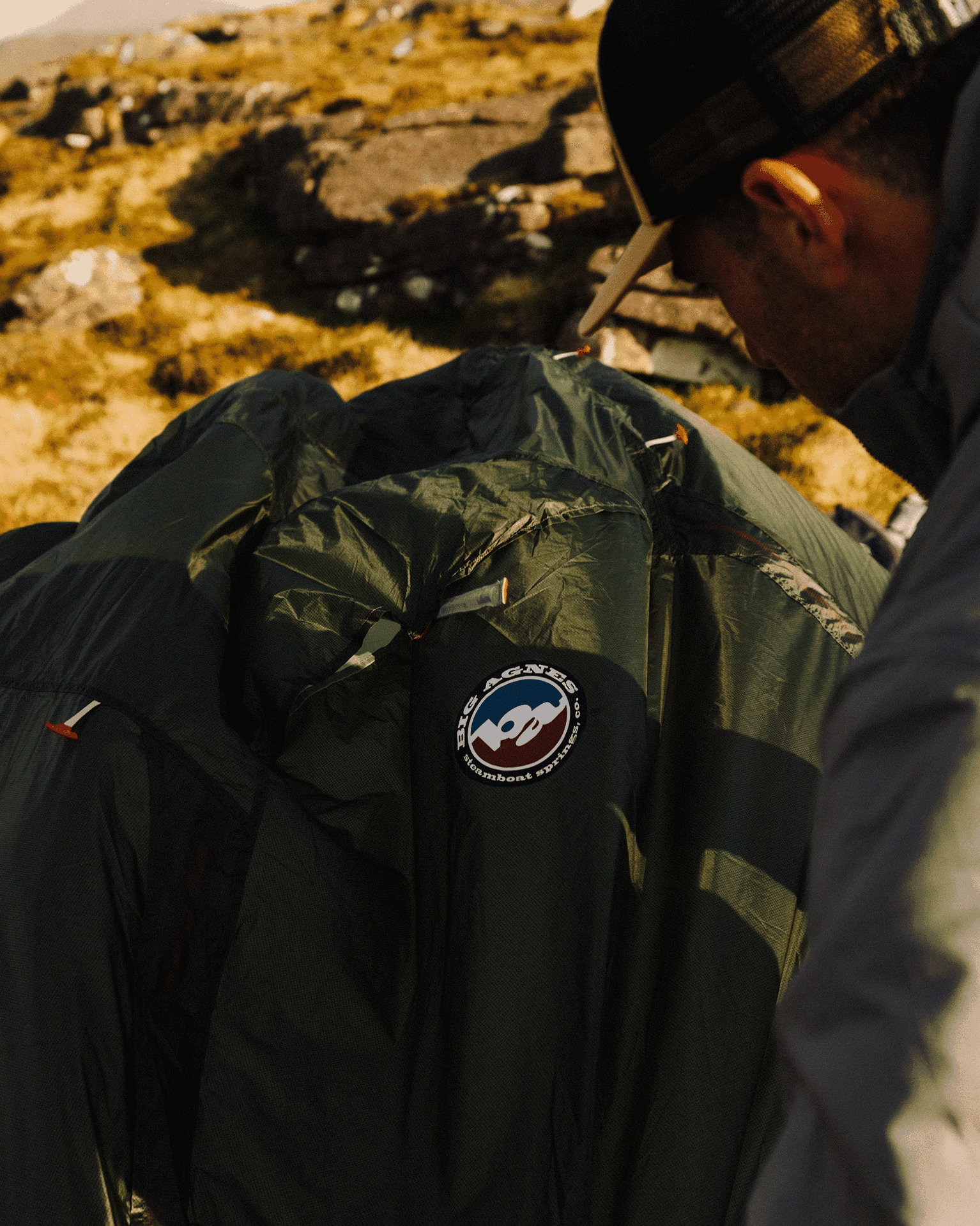 A person sets up a dark green Big Agnes tent on rocky terrain under sunlight, wearing a cap and jacket.
