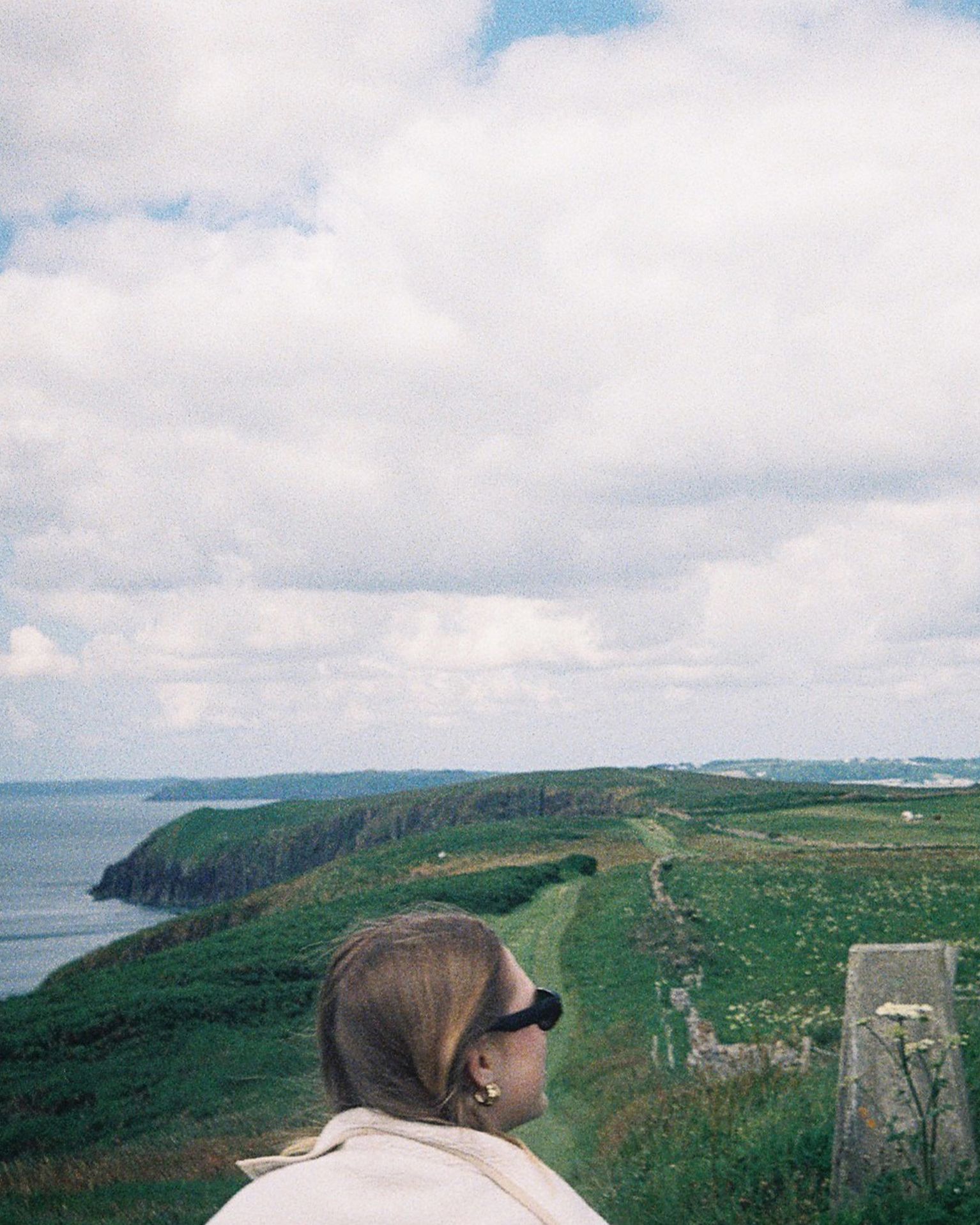 A person in sunglasses gazes at a scenic coastal landscape with green cliffs and a cloudy sky.