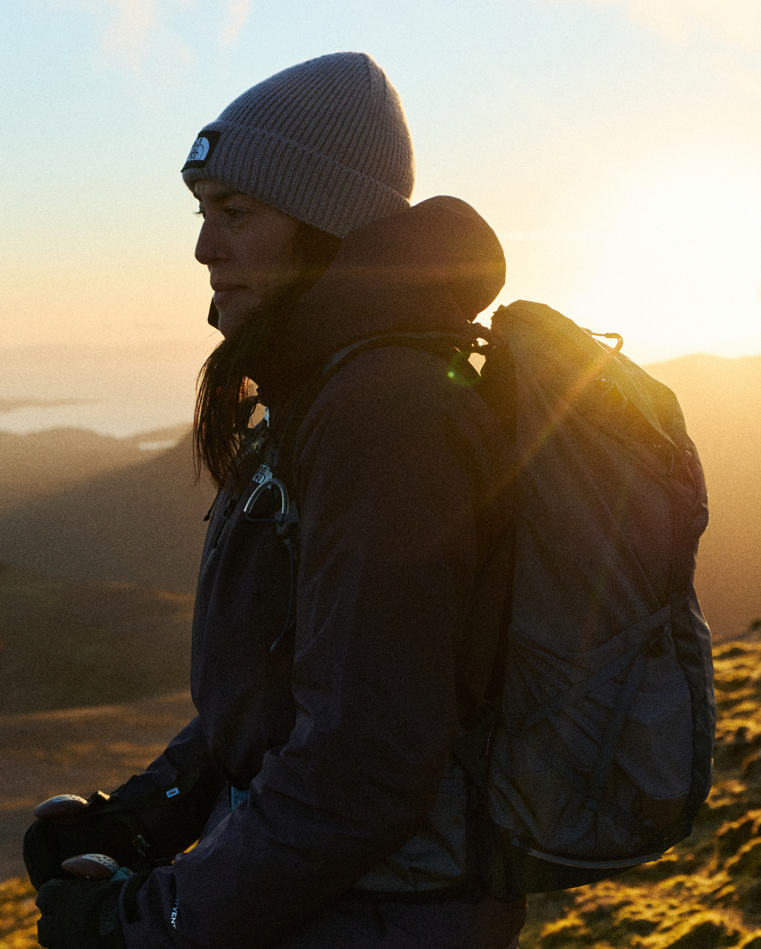 A person in mountain gear stands outdoors at sunrise, wearing a grey beanie and carrying a backpack, looking confidently ahead.