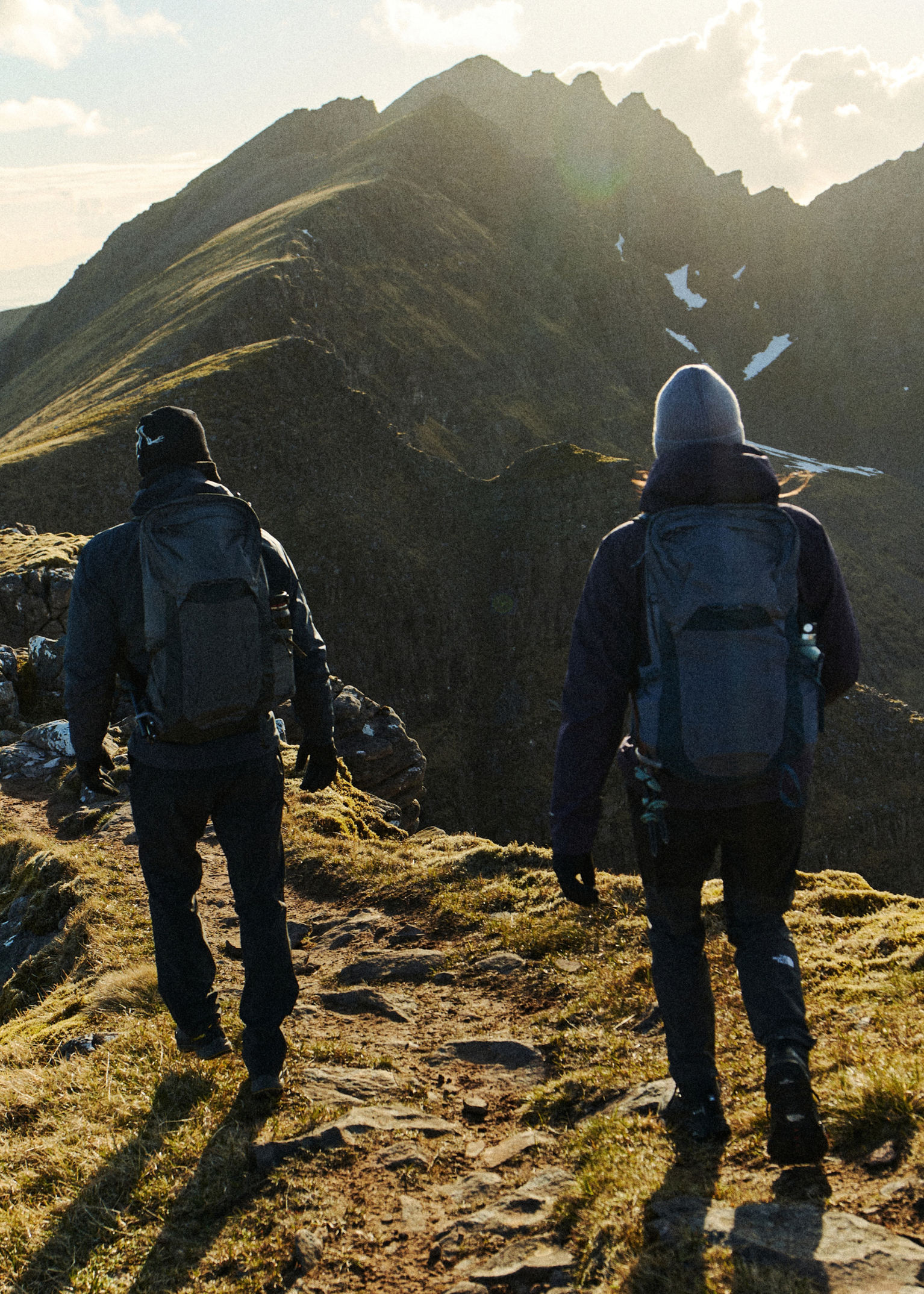 Two hikers with Osprey Talon and Tempest backpacks walking along a mountain ridge path, with rugged peaks and a clear sky in the background.