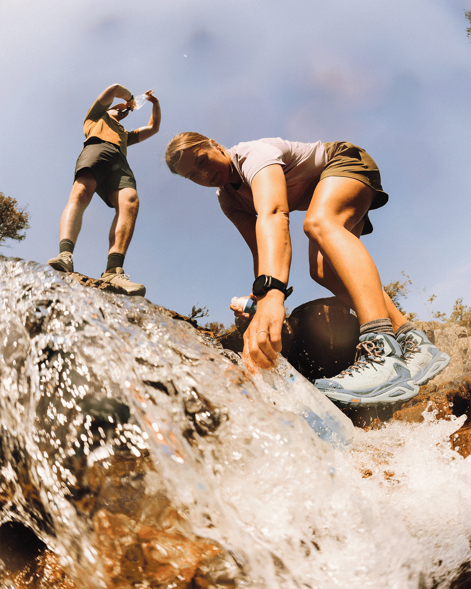 Two hikers collecting water from a stream in the mountains, wearing hiking clothes and sturdy boots.