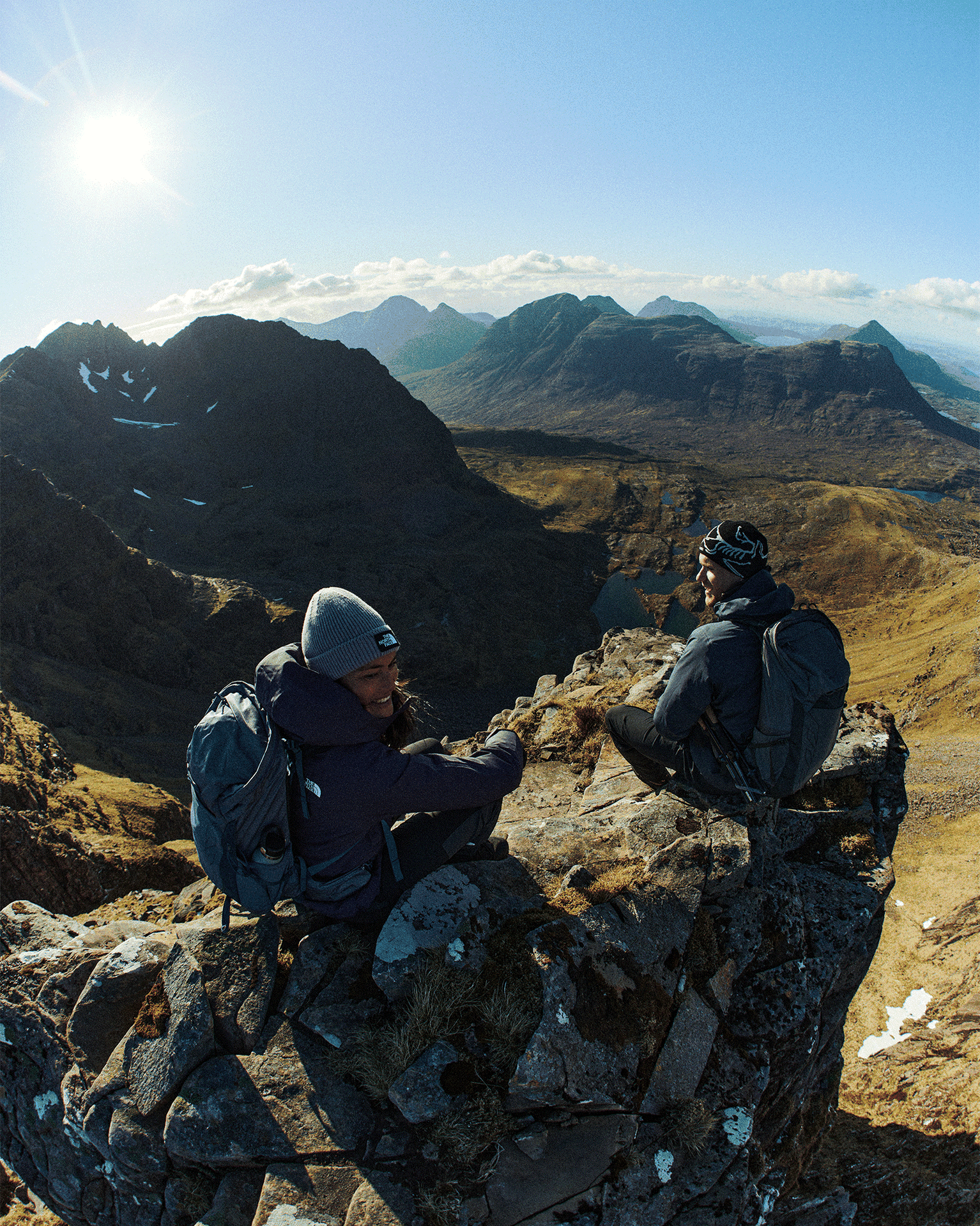 Two hikers in mountain gear sit on a rocky cliff with a stunning mountain landscape and bright sun in the background.