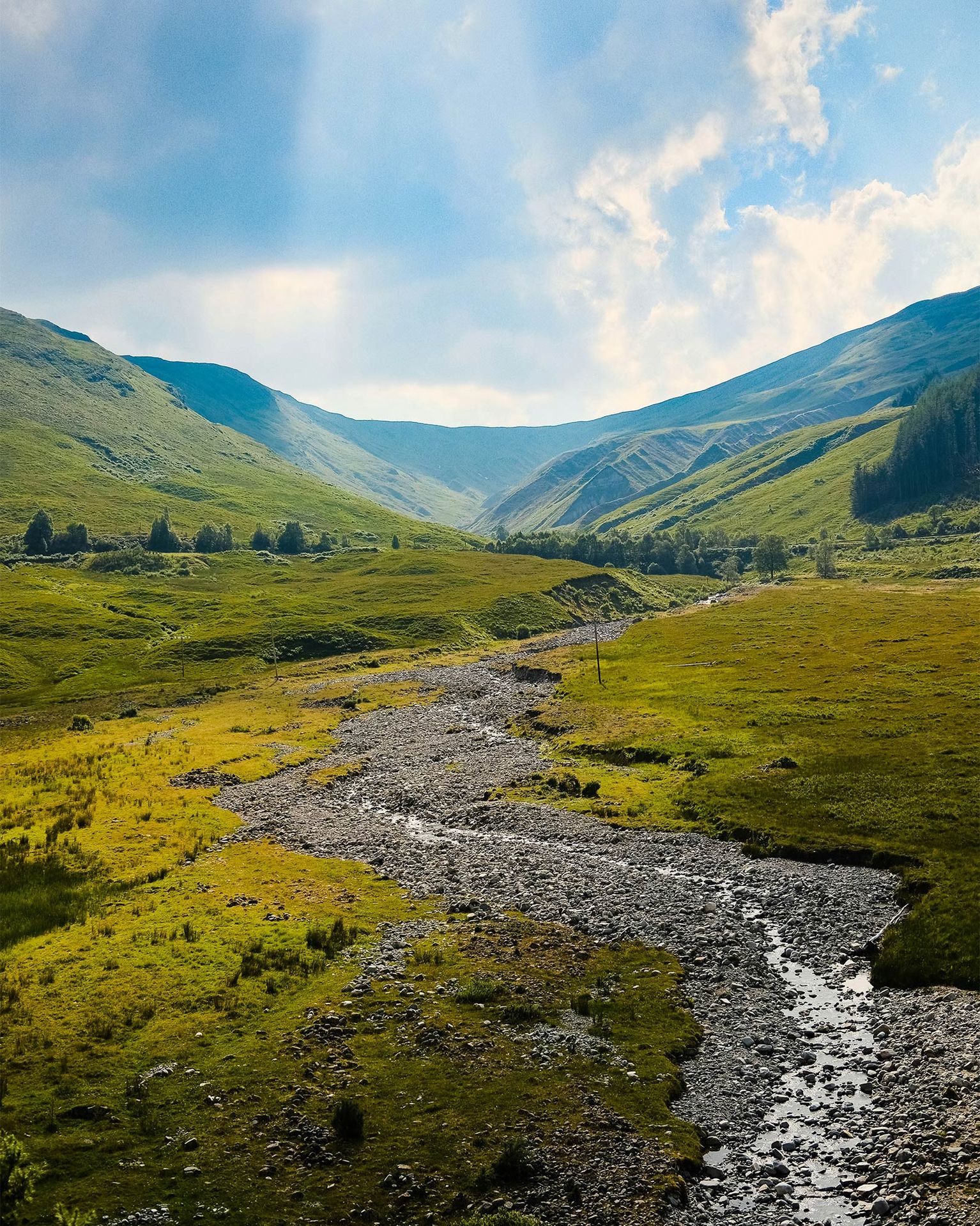 A scenic view of a rocky stream winding through a lush green valley under a clear blue sky with clouds.