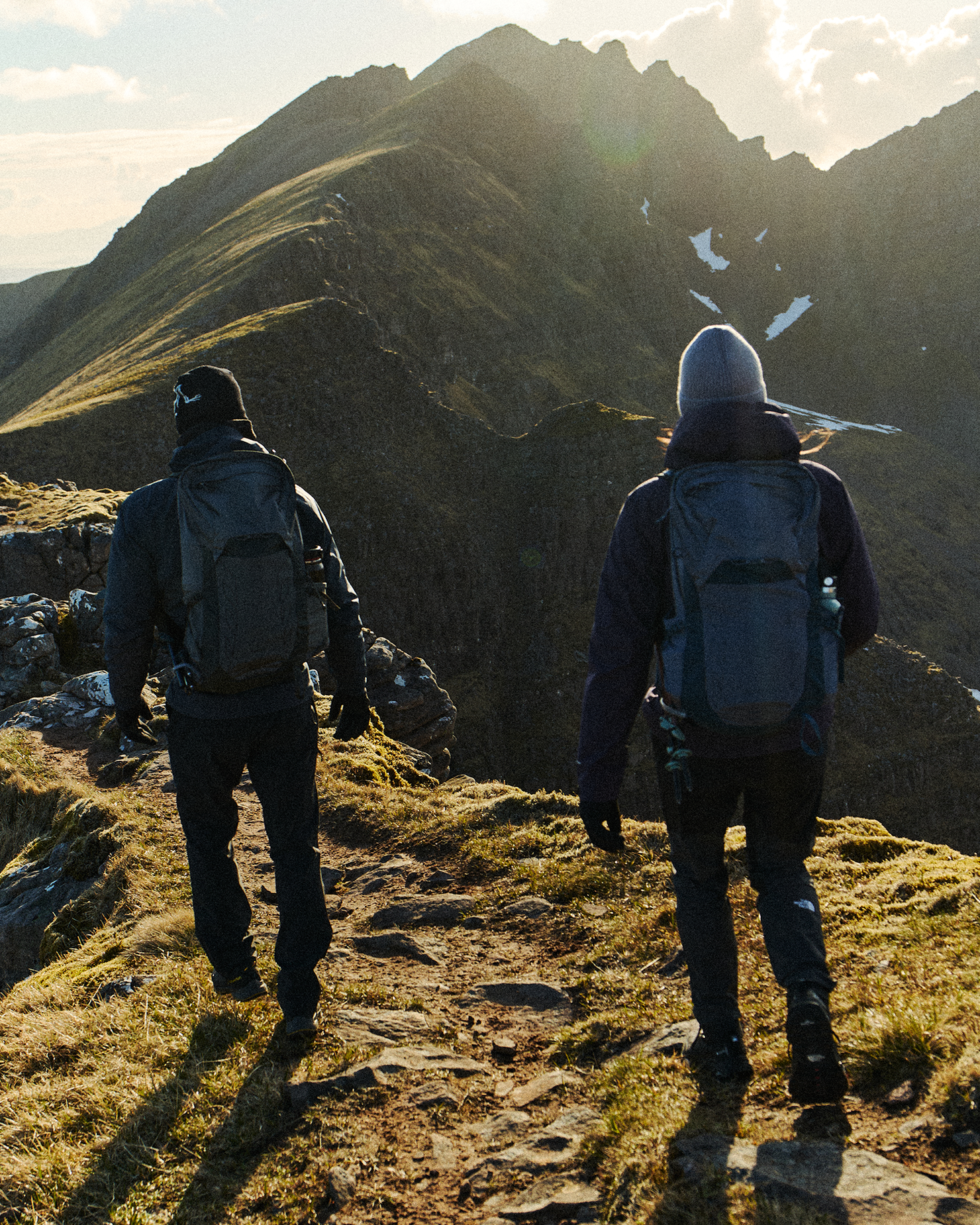 Two hikers with backpacks walk along a rugged mountain trail at sunrise, surrounded by stunning peaks and soft morning light.