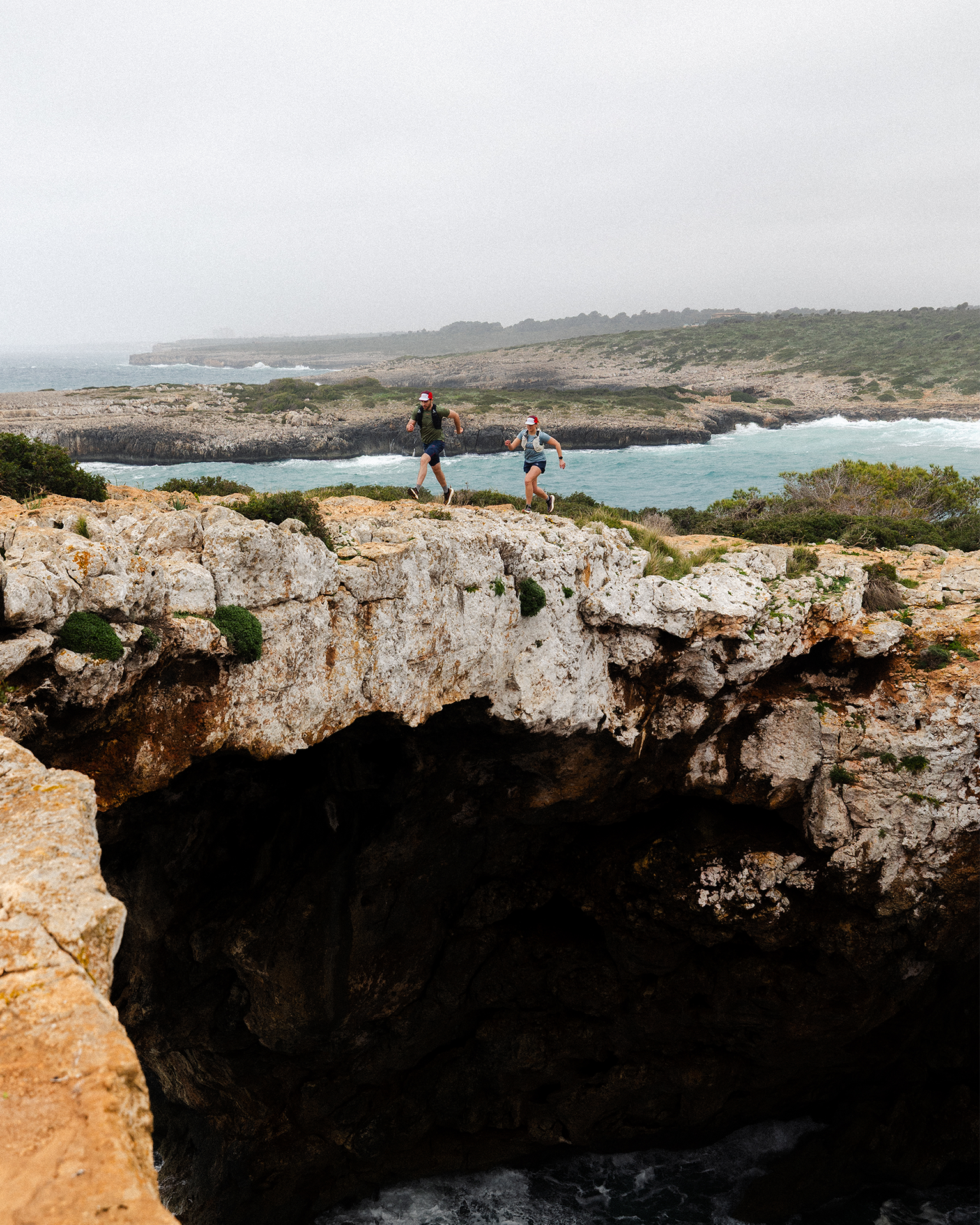 Two people trail running on rocky coastal cliffs with a scenic ocean and rugged landscape in the background.