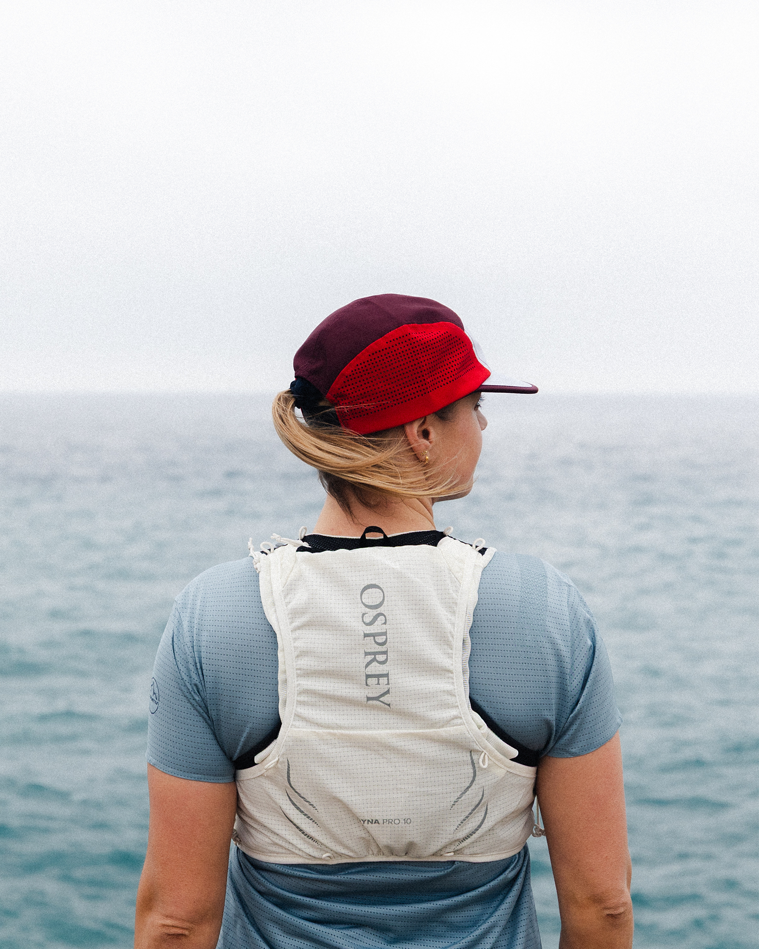 A person wearing a red hat and white Osprey backpack faces the ocean, dressed in a blue top.