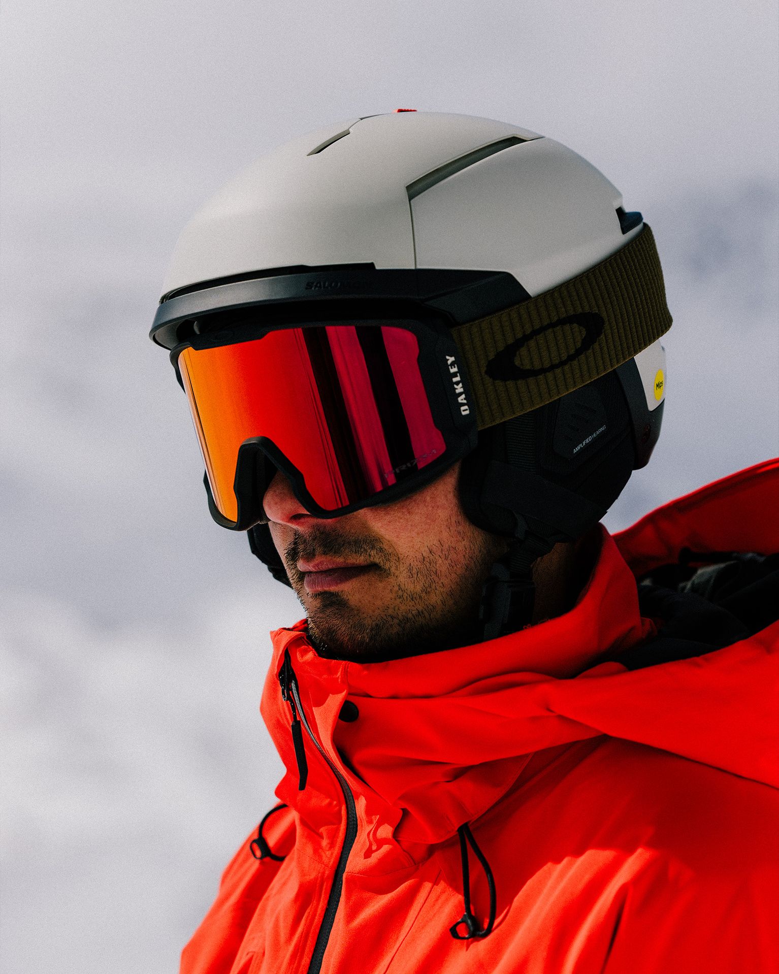 Person in a red jacket wearing a white helmet and reflective goggles, standing against a snowy background.