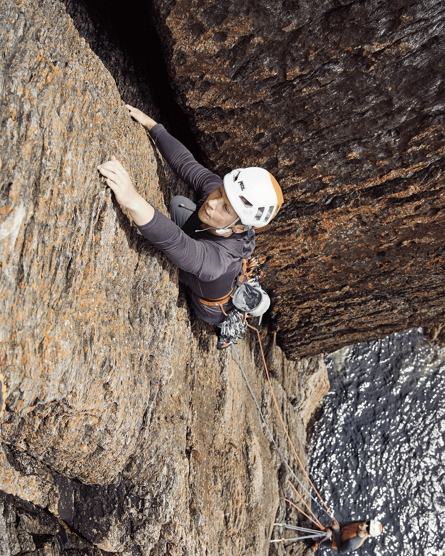 Climber with helmet and gear scales a rugged rock face above water, demonstrating skill and focus.