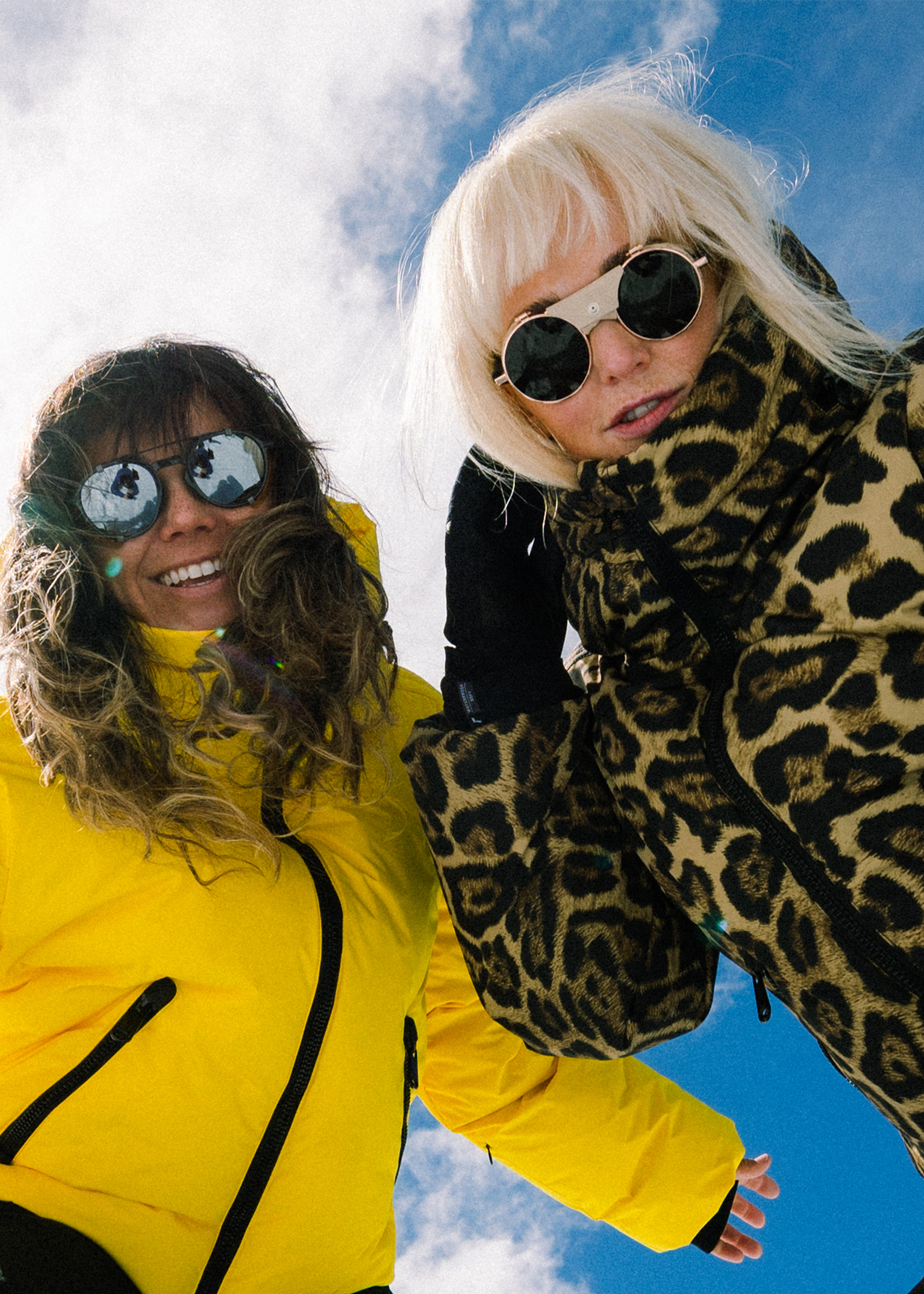 Low-angle shot of two people in mountain-sports outerwear—one bright yellow jacket, one leopard-print parka—wearing sunglasses against a clear blue sky.