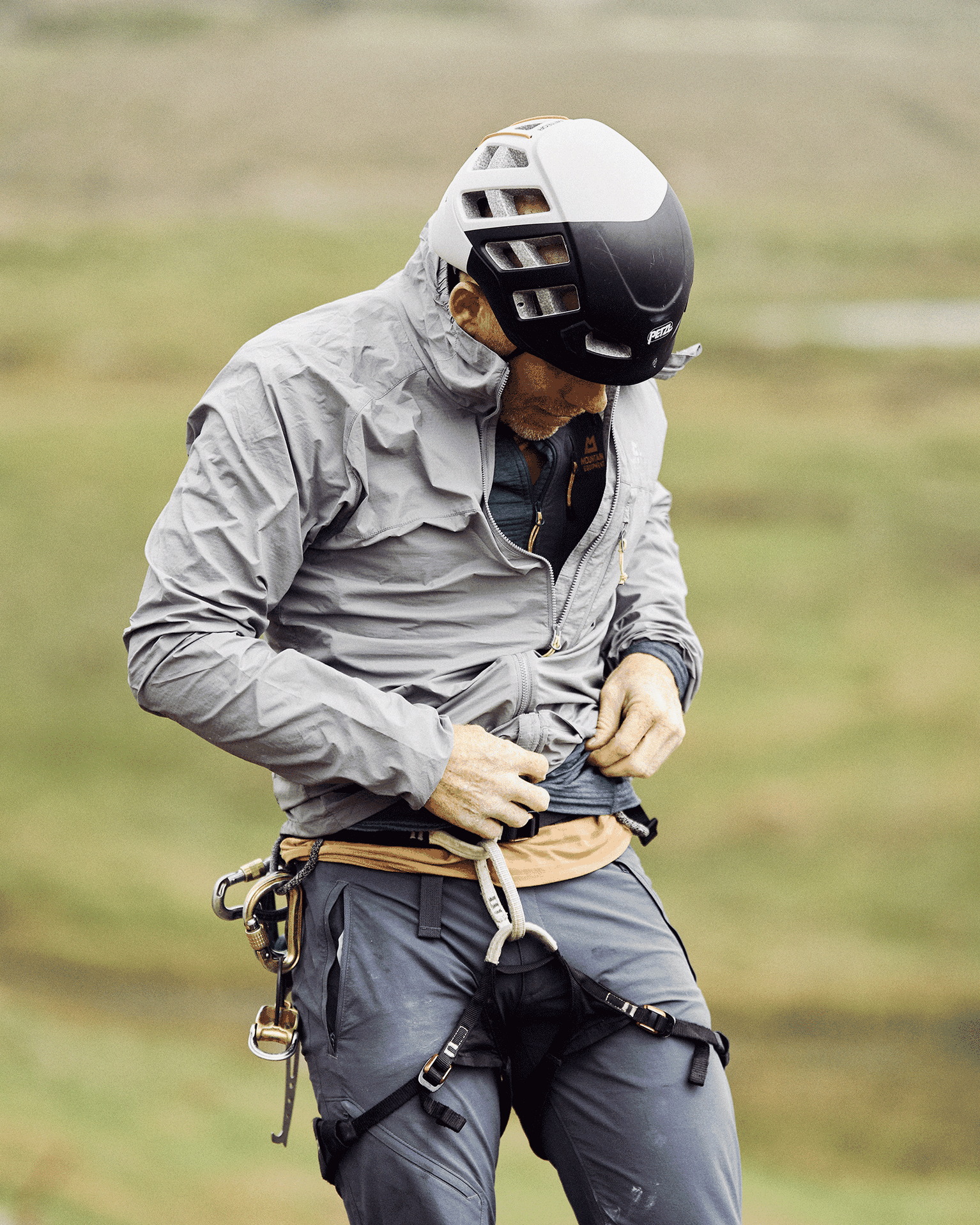 Man wearing a helmet, grey jacket, and climbing harness, adjusting gear outdoors.