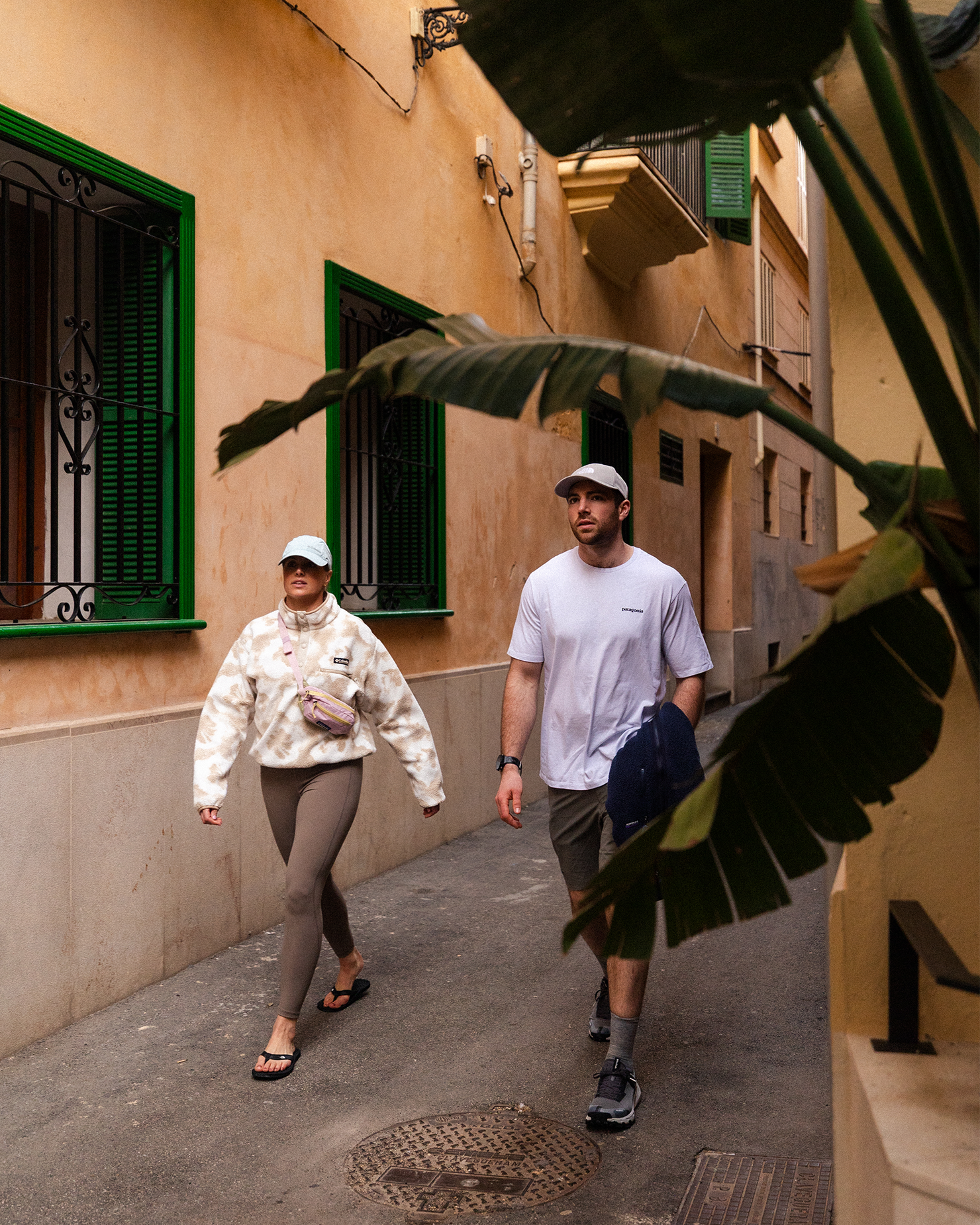Two people in casual clothing and caps walking down a narrow street, partially obscured by a large plant in the foreground.