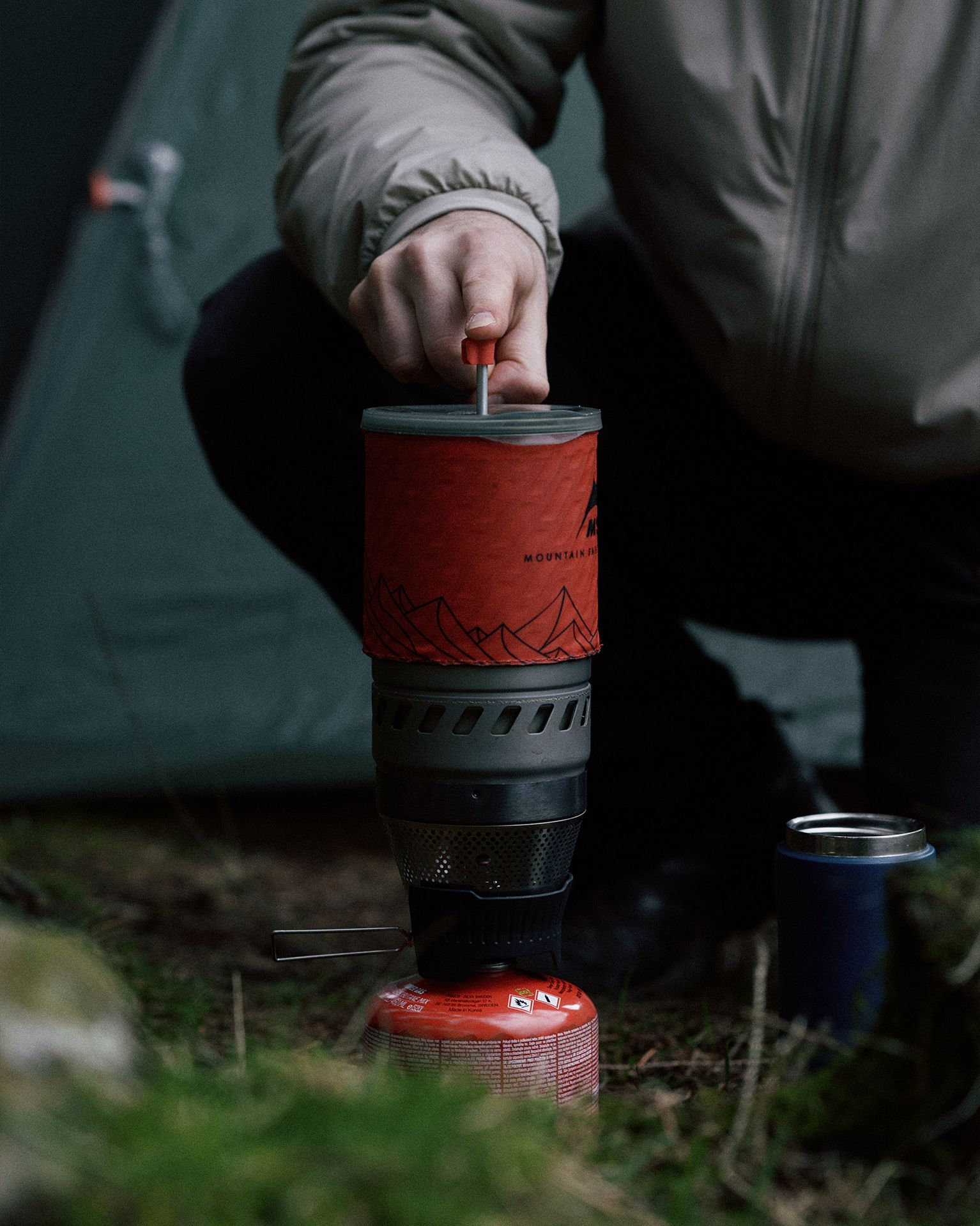 Person using a portable camping stove with a coffee press attachment outside a tent in a wooded area.
