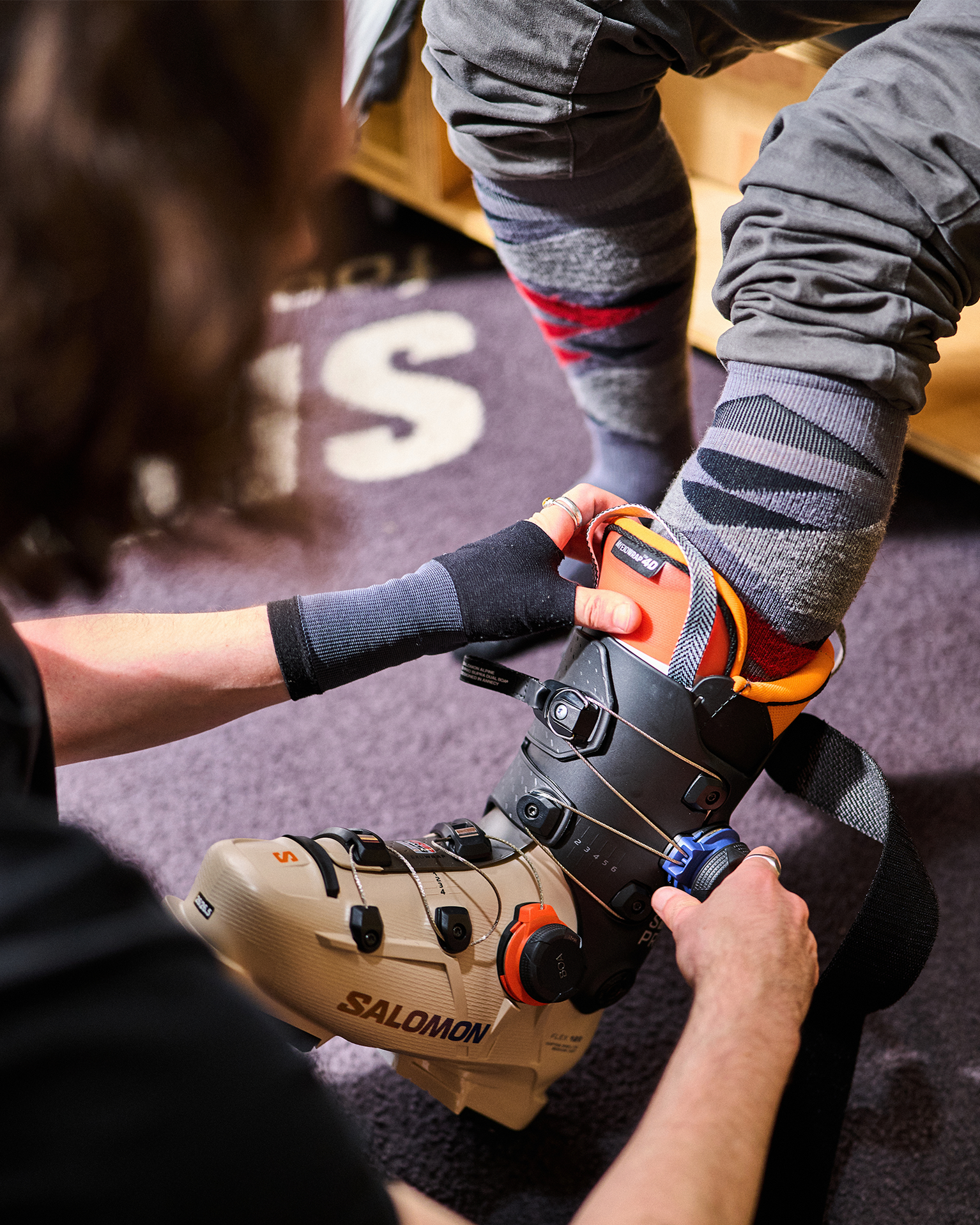 Person helping another adjust a beige ski boot with black straps, indoors. The person being helped is wearing gray pants and colorful socks.