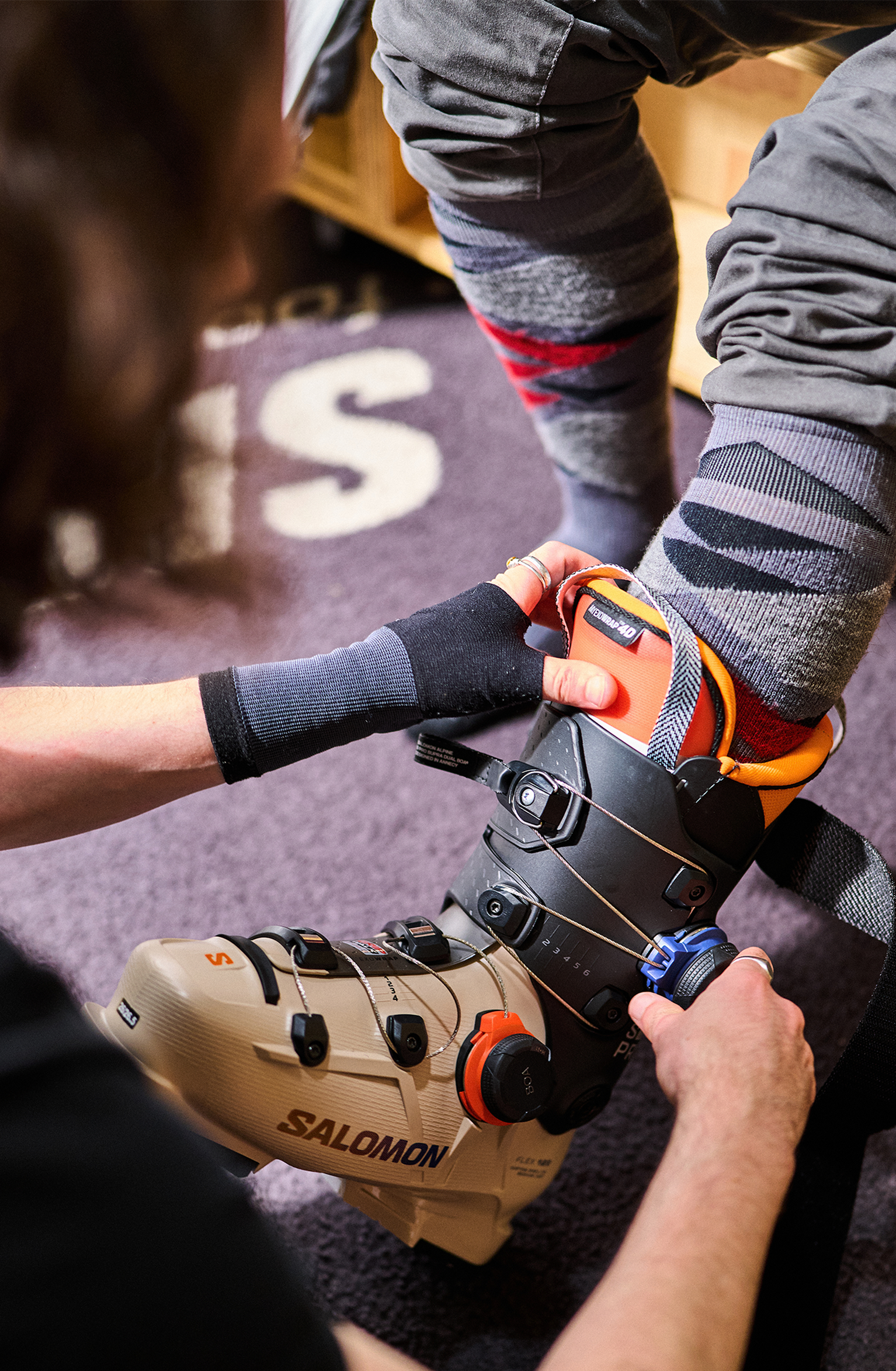 Person helping another adjust a beige ski boot with black straps, indoors. The person being helped is wearing gray pants and colorful socks.