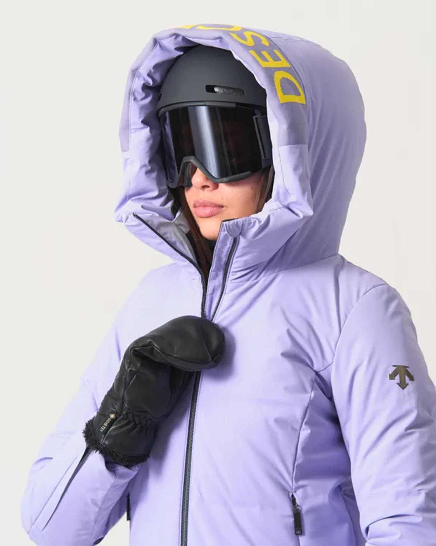 Person in lilac ski jacket with hood up, black helmet and goggles, wearing leather mitts.