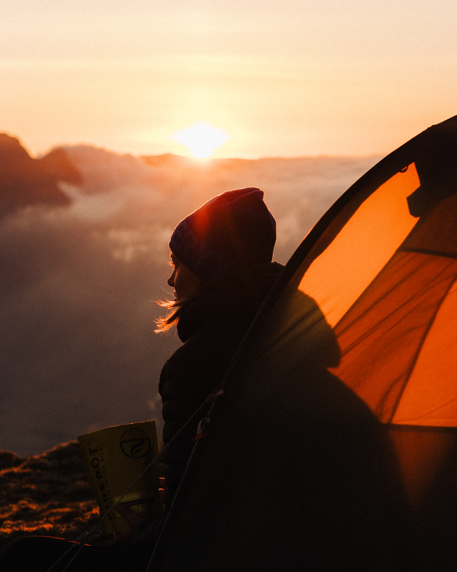 Person sitting by a glowing tent, facing a sunrise over misty mountains, wearing a beanie and holding a cup.