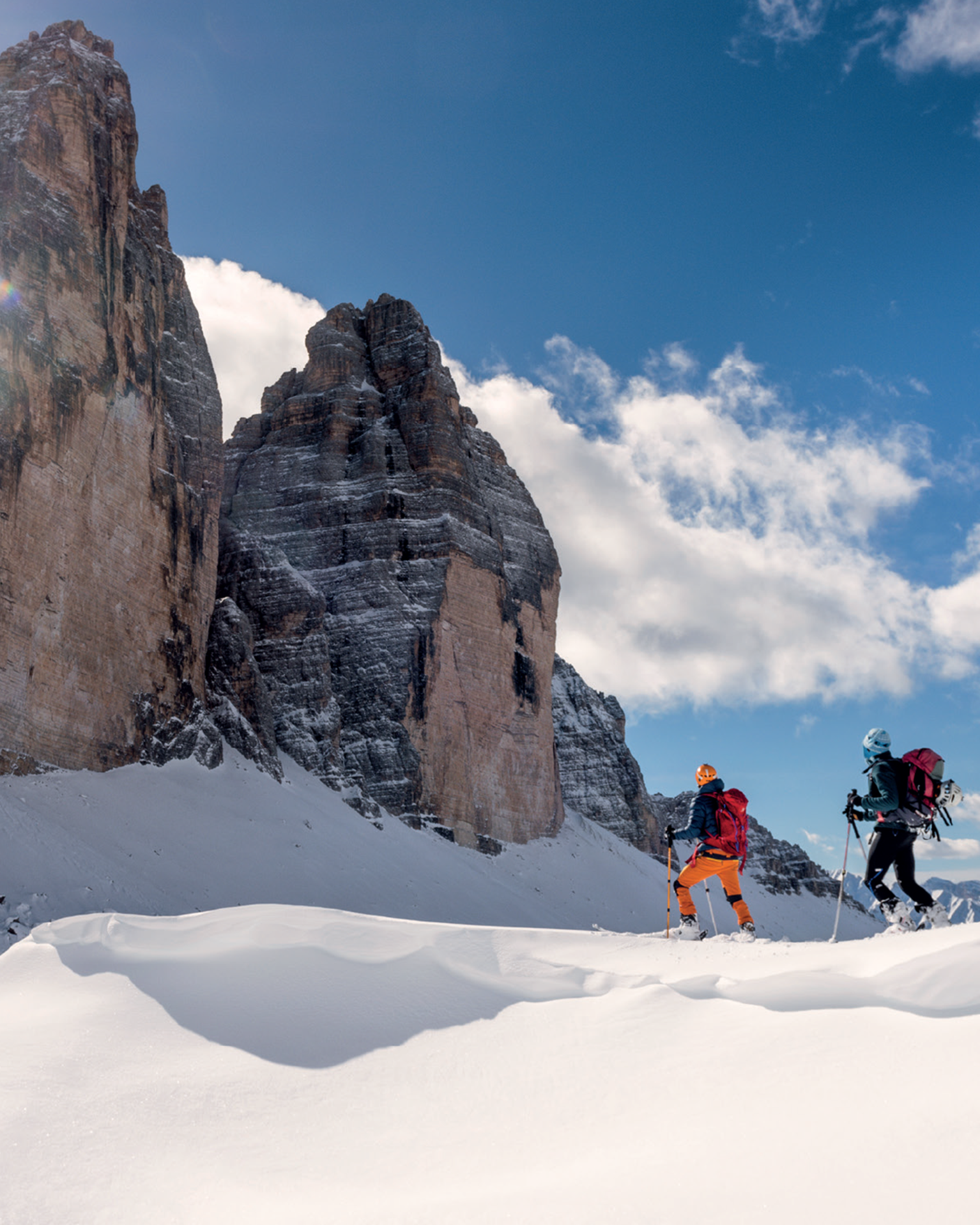 Two skiers traverse snowy terrain, with towering mountain peaks under a clear blue sky.