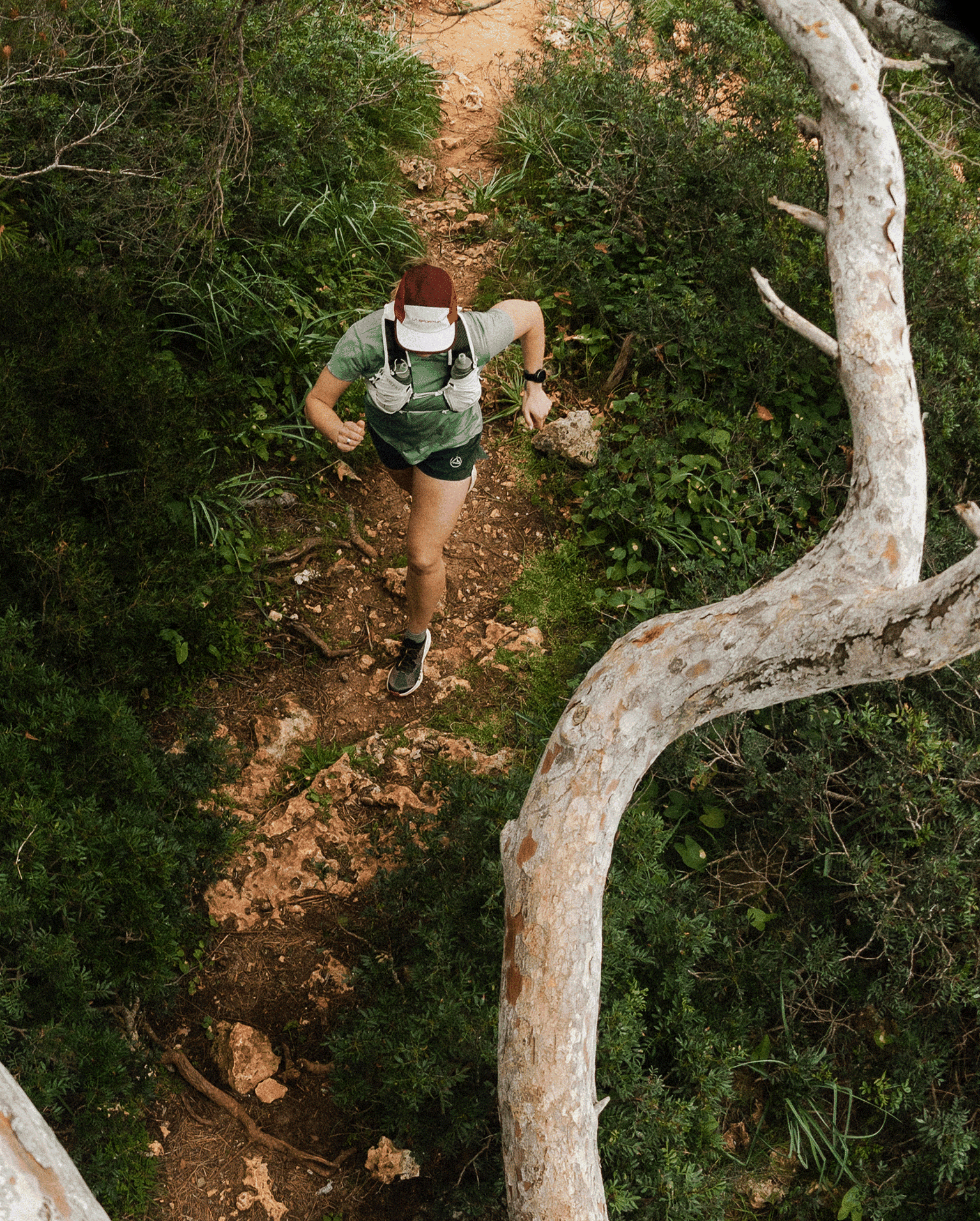 Trail runner wearing a cap and hydration pack navigates a rocky forest path surrounded by lush greenery, viewed from above.