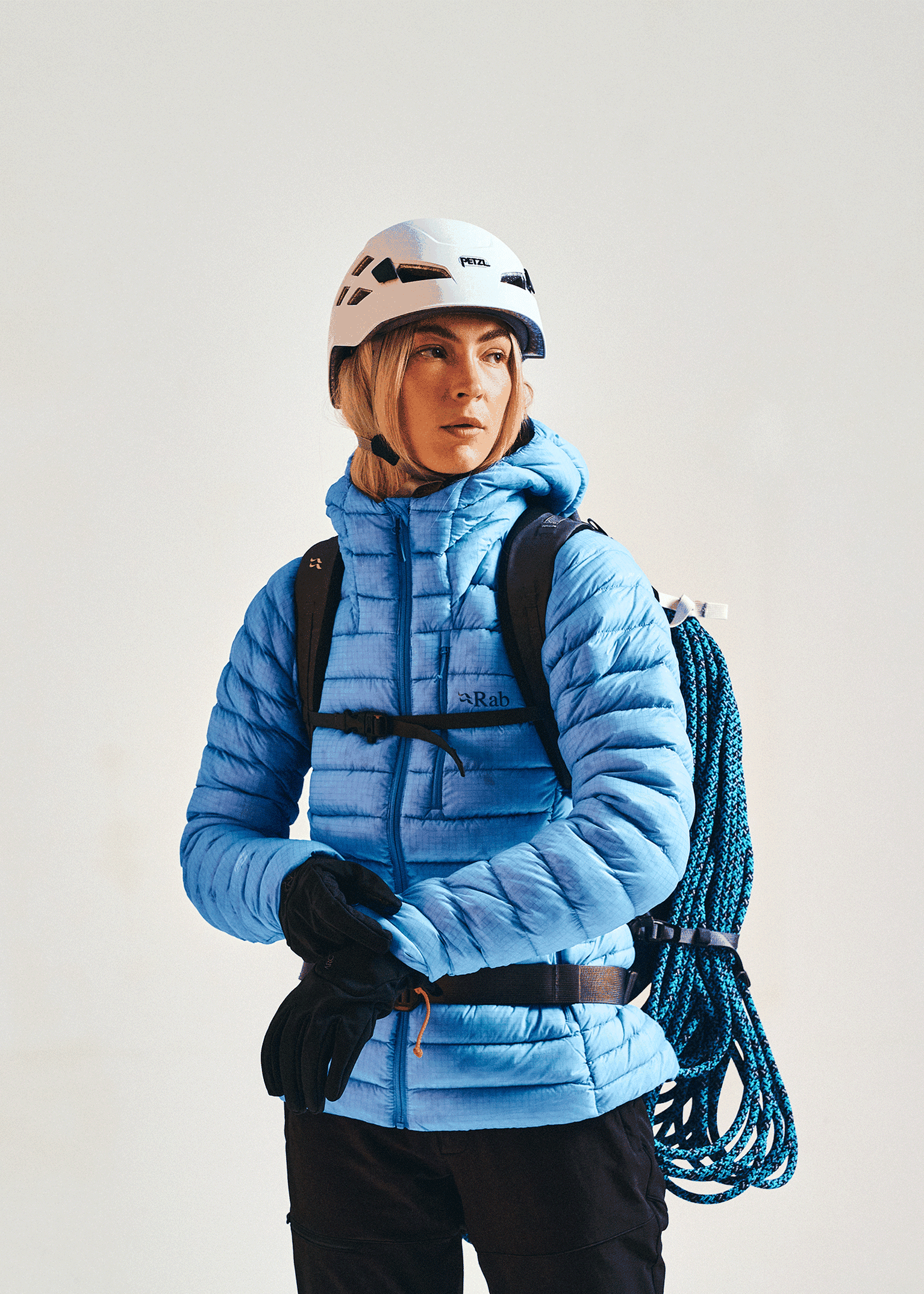 A person wearing a blue Rab Microlight X jacket, white helmet, black gloves, and a backpack with climbing rope.