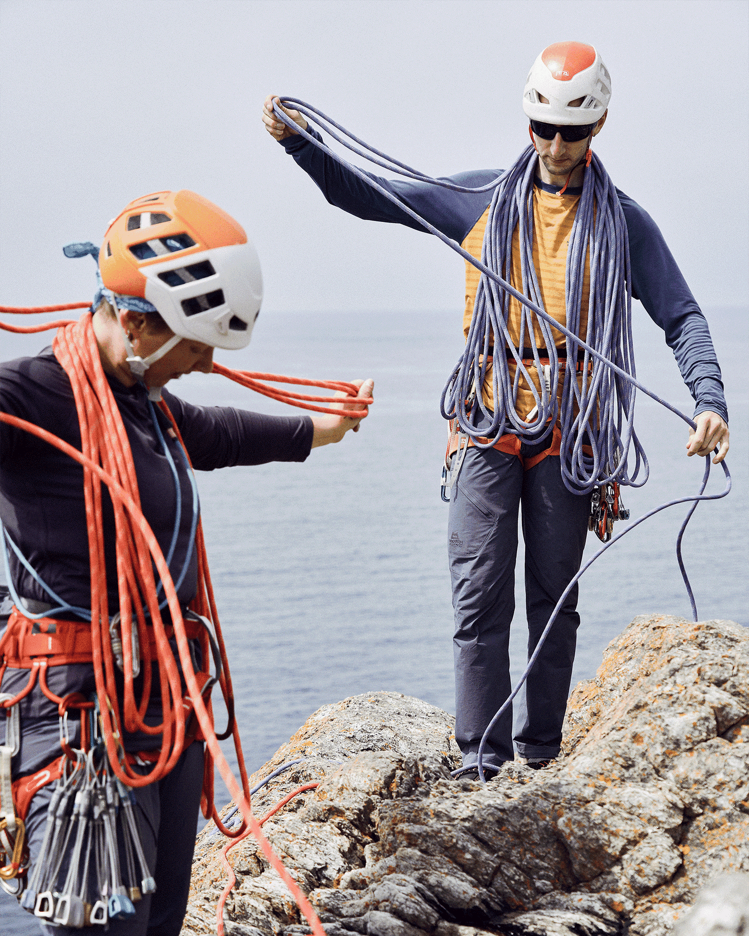 Two climbers in helmets and gear organise ropes on a rocky cliff overlooking the sea, preparing for a climb.