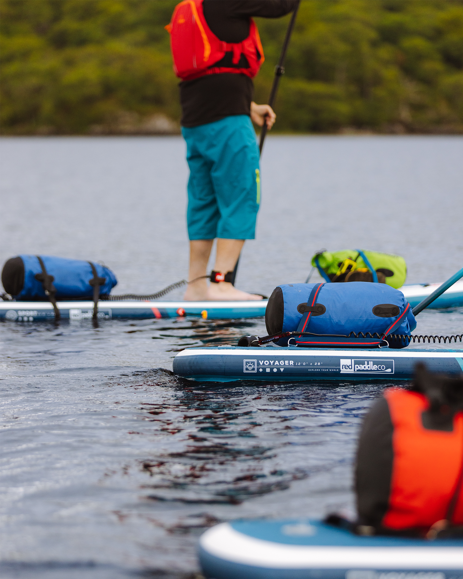 Person paddleboarding on calm water with gear bags, wearing a red life jacket and turquoise shorts. Forested shoreline in the background.