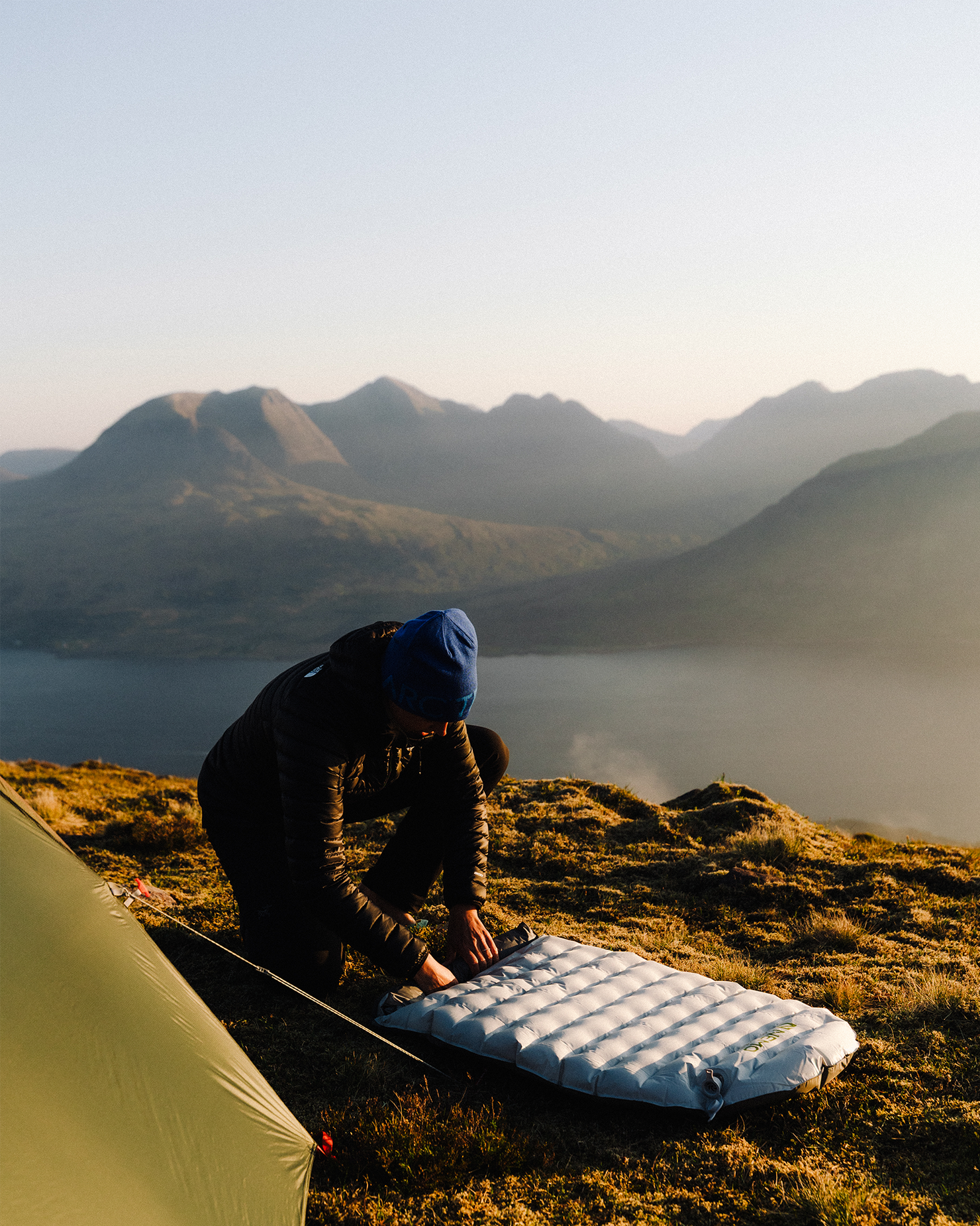 Camper setting up an inflatable mattress on a hillside, with a tent nearby and mountains in the background at sunrise.