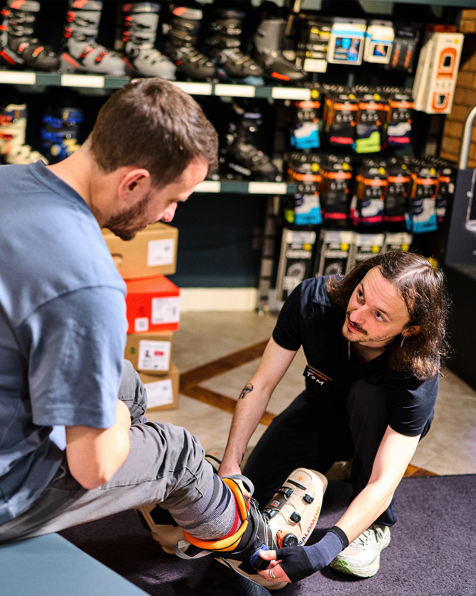 A store employee assists a customer with trying on ski boots in a shop, surrounded by shelves of ski gear and accessories.