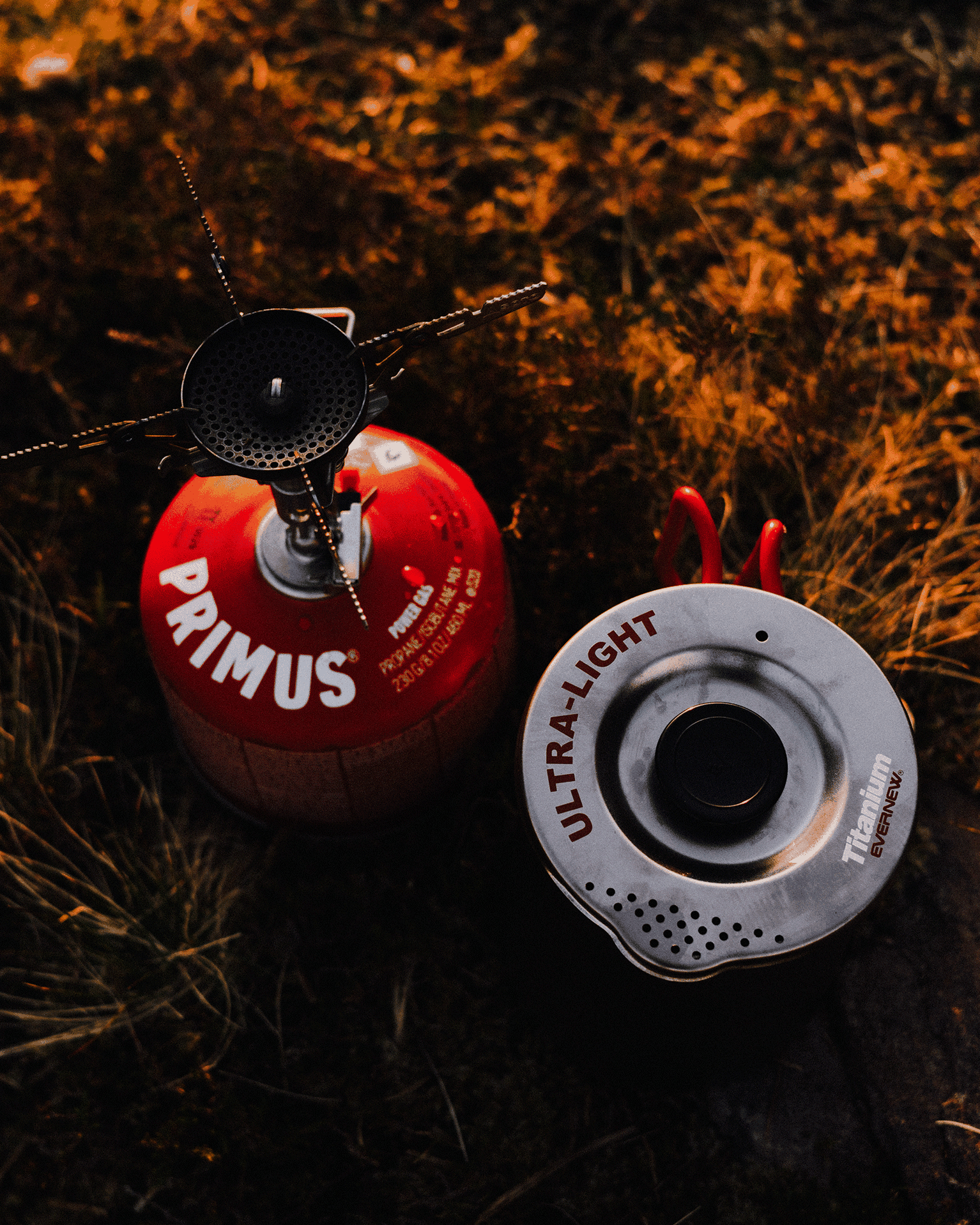 Camping stove and red fuel canister on dry grass, illuminated by warm light, with "Primus" and "Ultra-Light" visible on the canisters.
