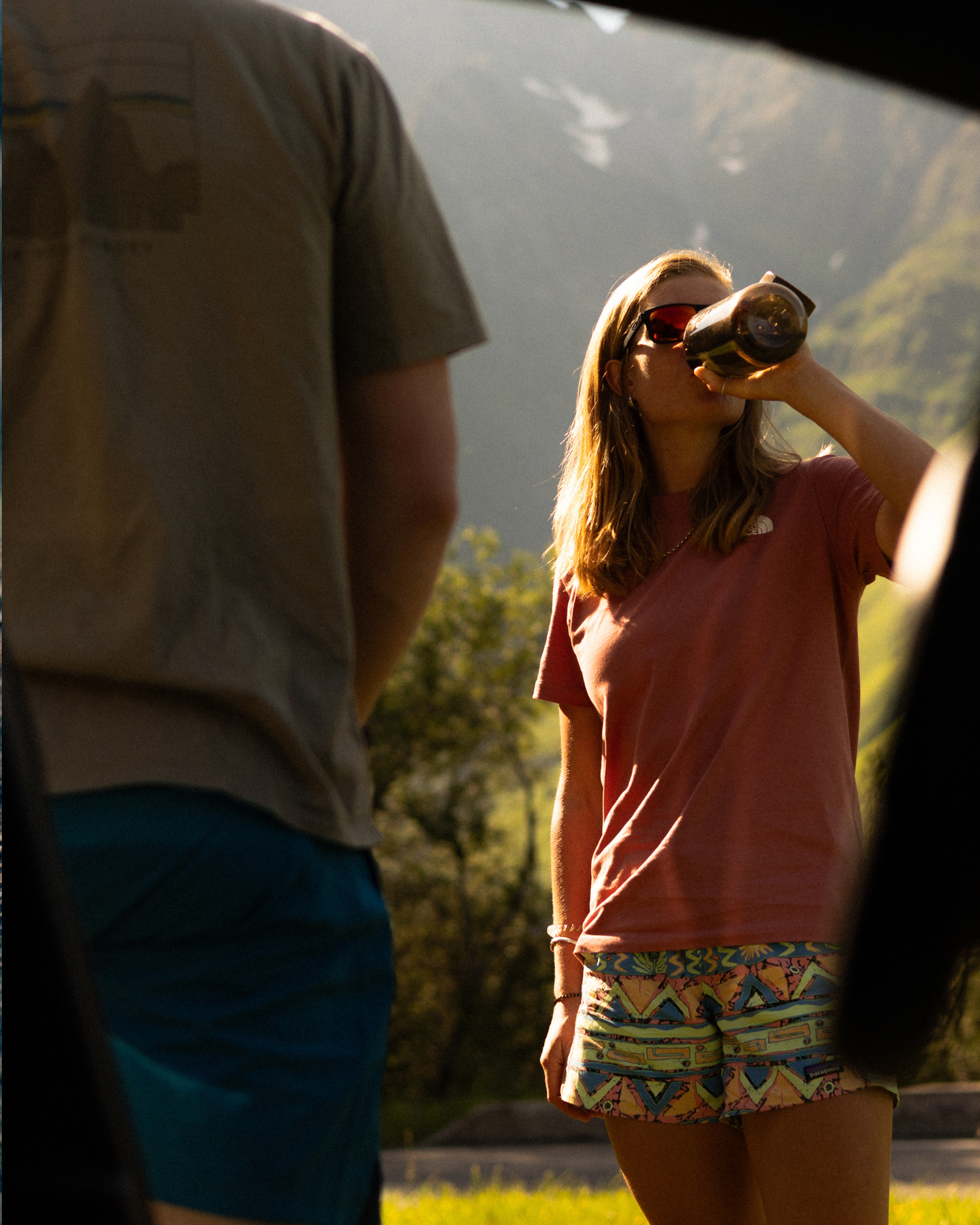 Woman in sunglasses drinks from a bottle outdoors, wearing a pink shirt and patterned shorts, with a person in the foreground and mountains behind.