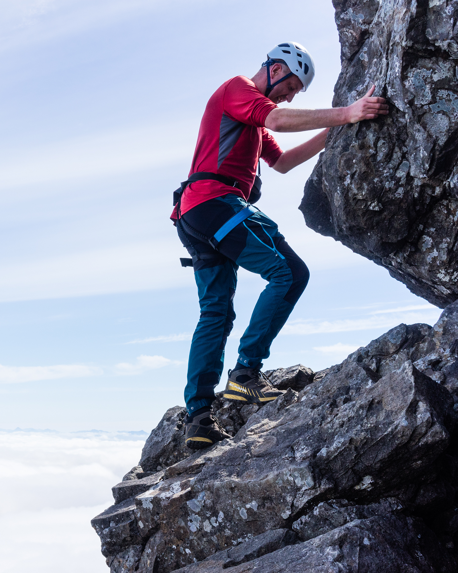 Climber wearing a red shirt, helmet, and harness, scaling a rocky mountain with a cloud-filled sky in the background.