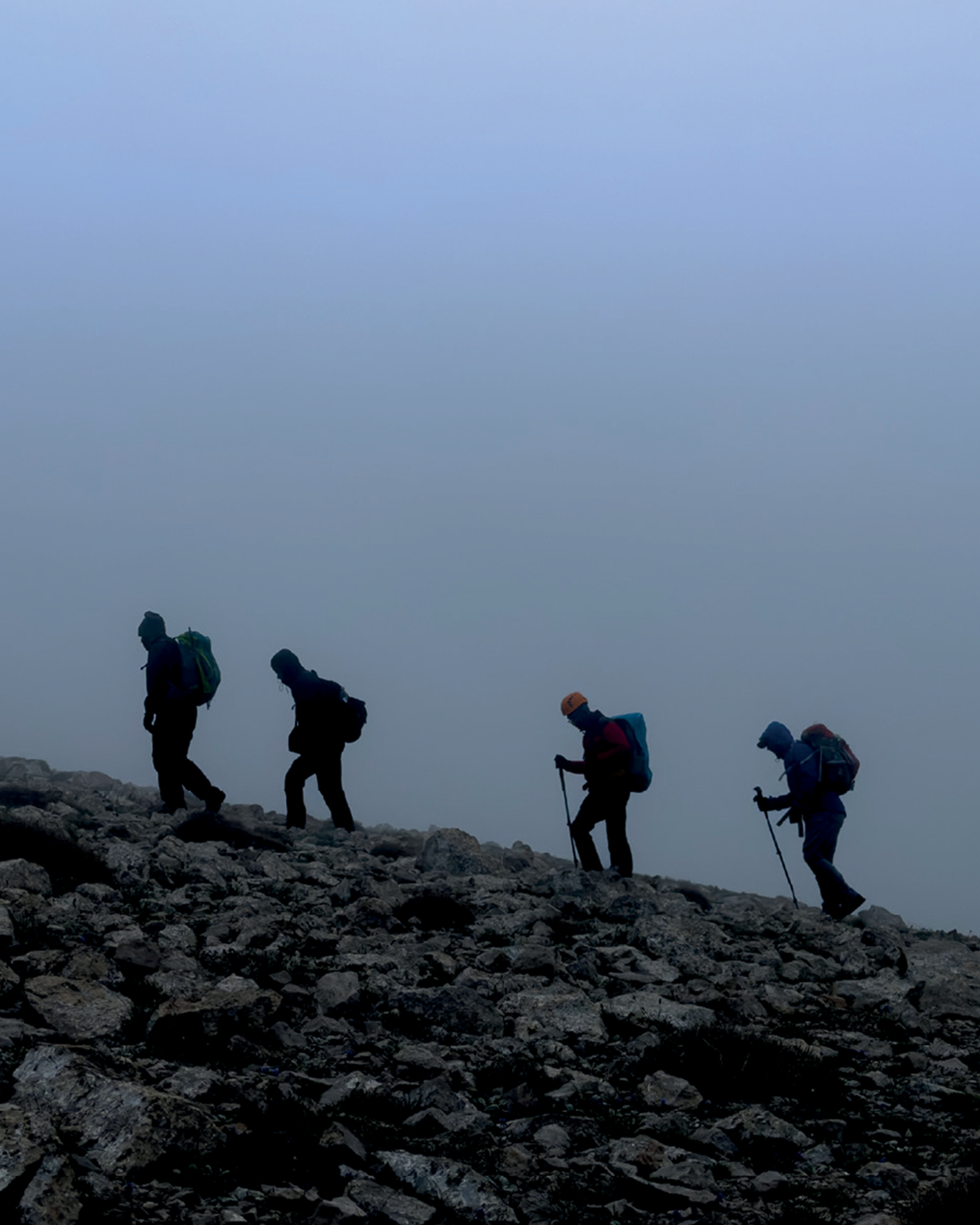 Four hikers, equipped with backpacks and trekking poles, ascend a rocky mountain slope on a foggy day.