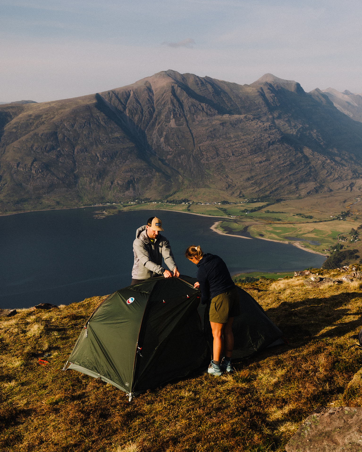 Two people set up a green tent on a hillside with a stunning view of mountains and a lake in the background.
