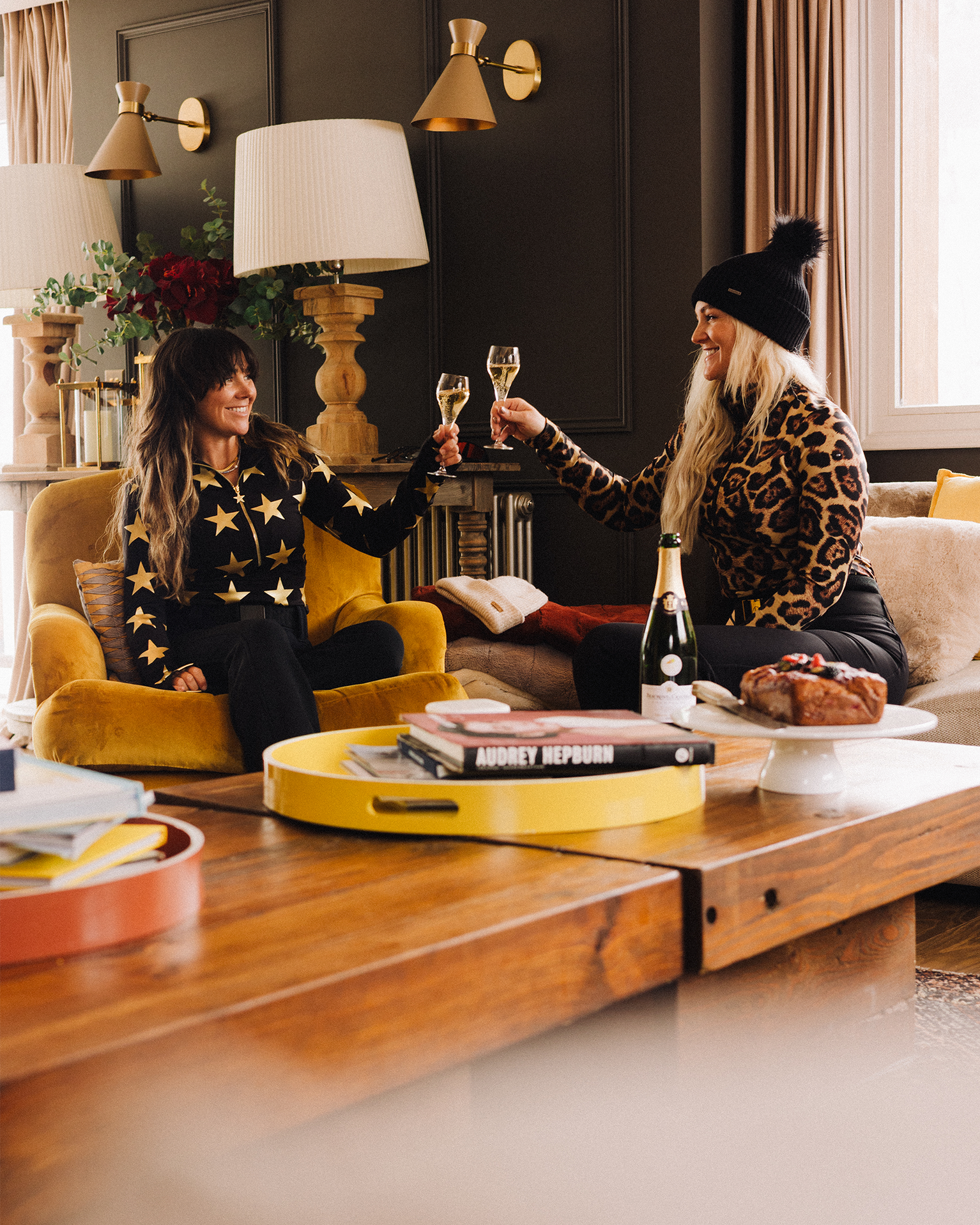 Two women in stylish outfits toast with champagne glasses in a cozy, warmly lit room with books and a cake on the table.