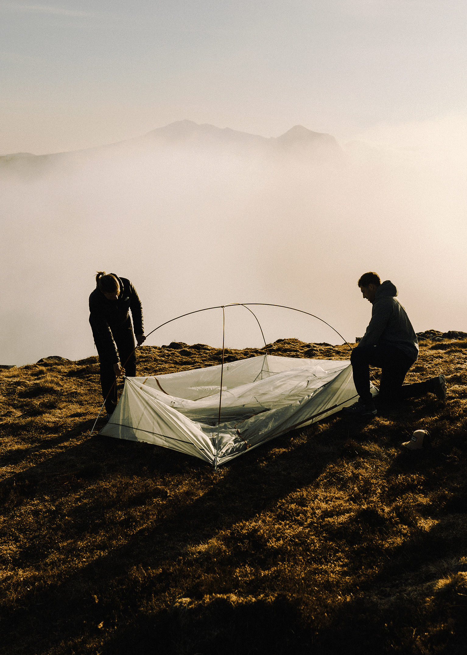 Two people setting up a tent on a grassy mountain slope, with a misty backdrop of hills and a soft sky.