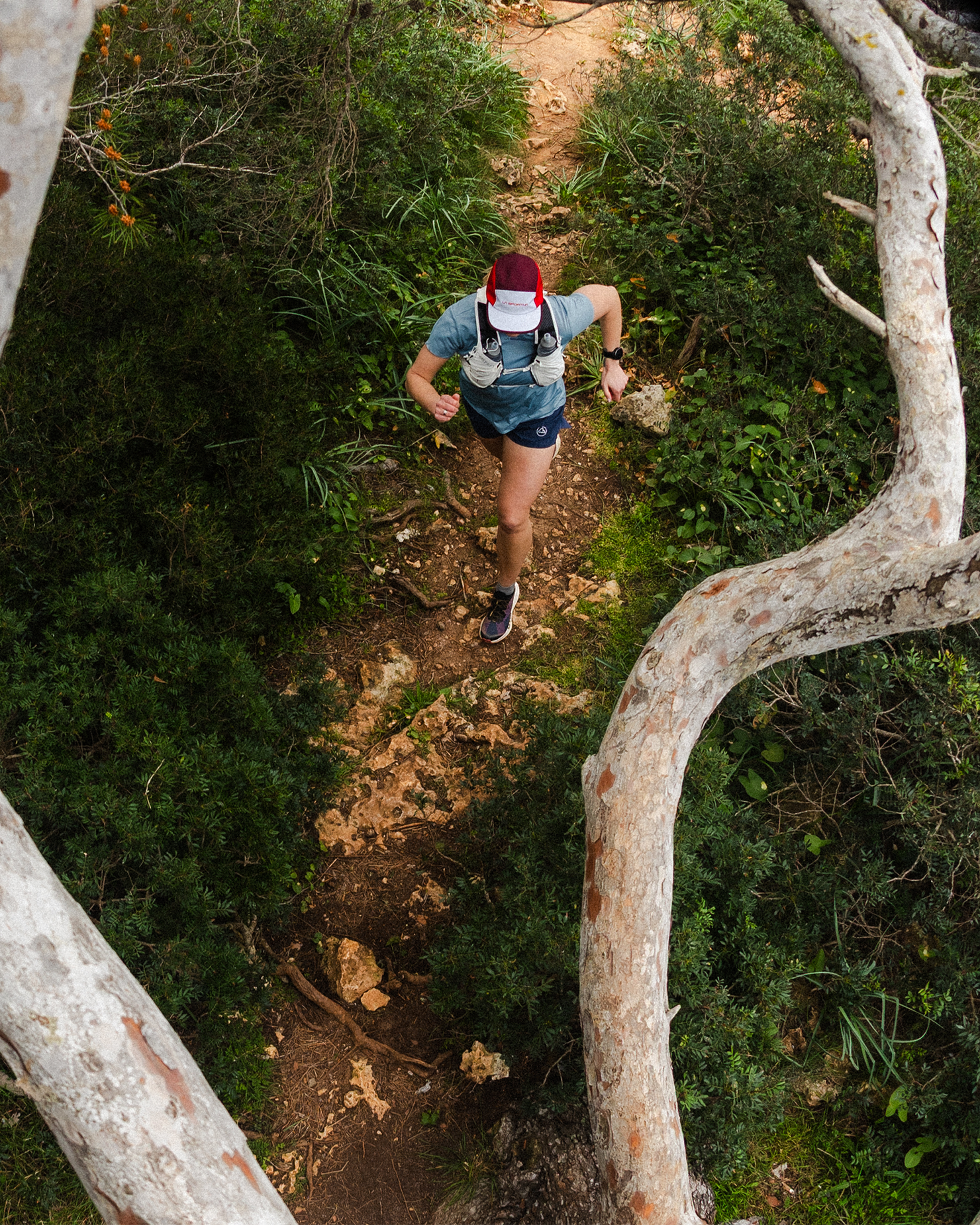 Trail runner navigating a narrow, rocky path through dense forest, viewed from above between tree branches.