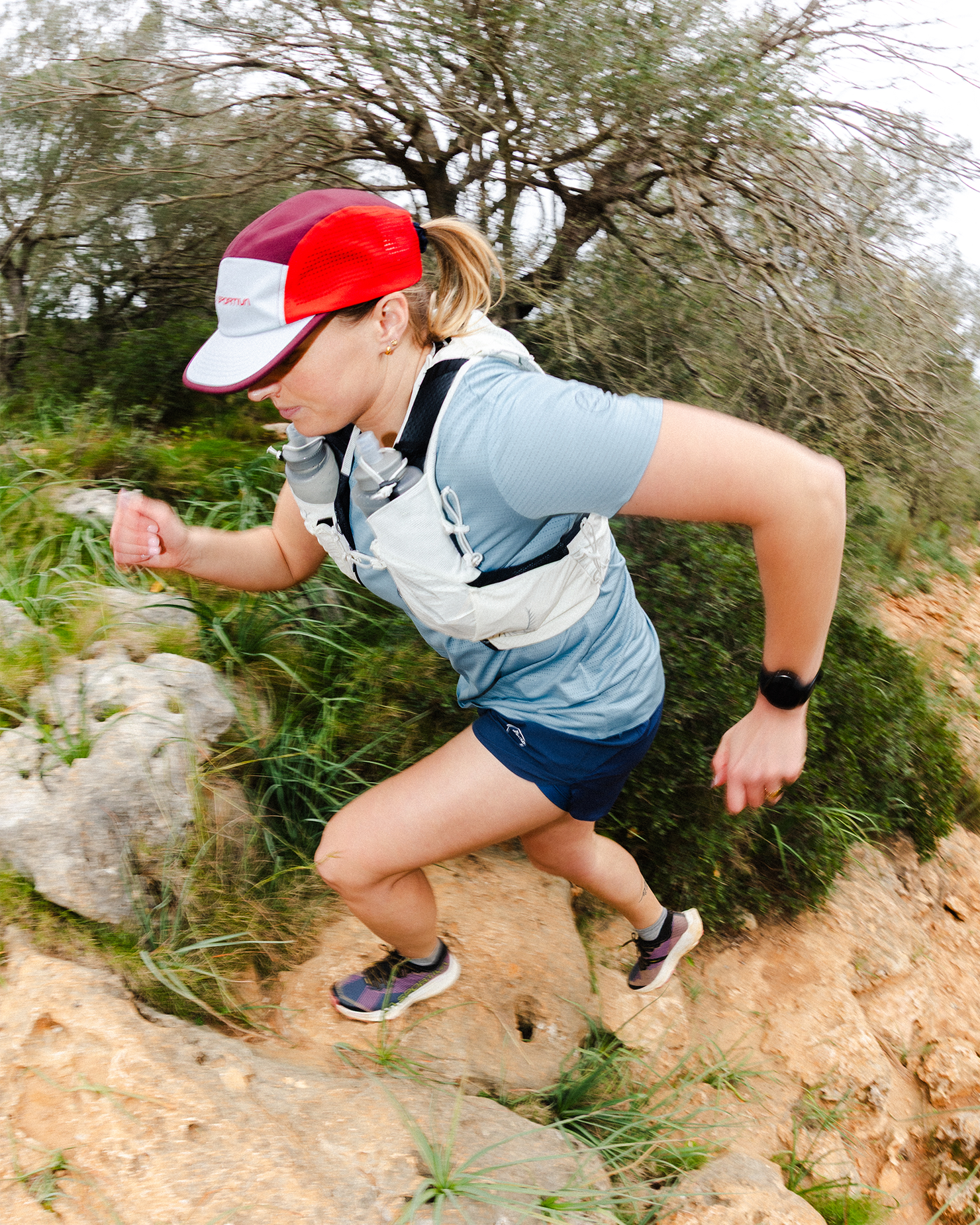 Woman trail running uphill on a rocky path, wearing a blue shirt, red cap, and vest, surrounded by greenery.