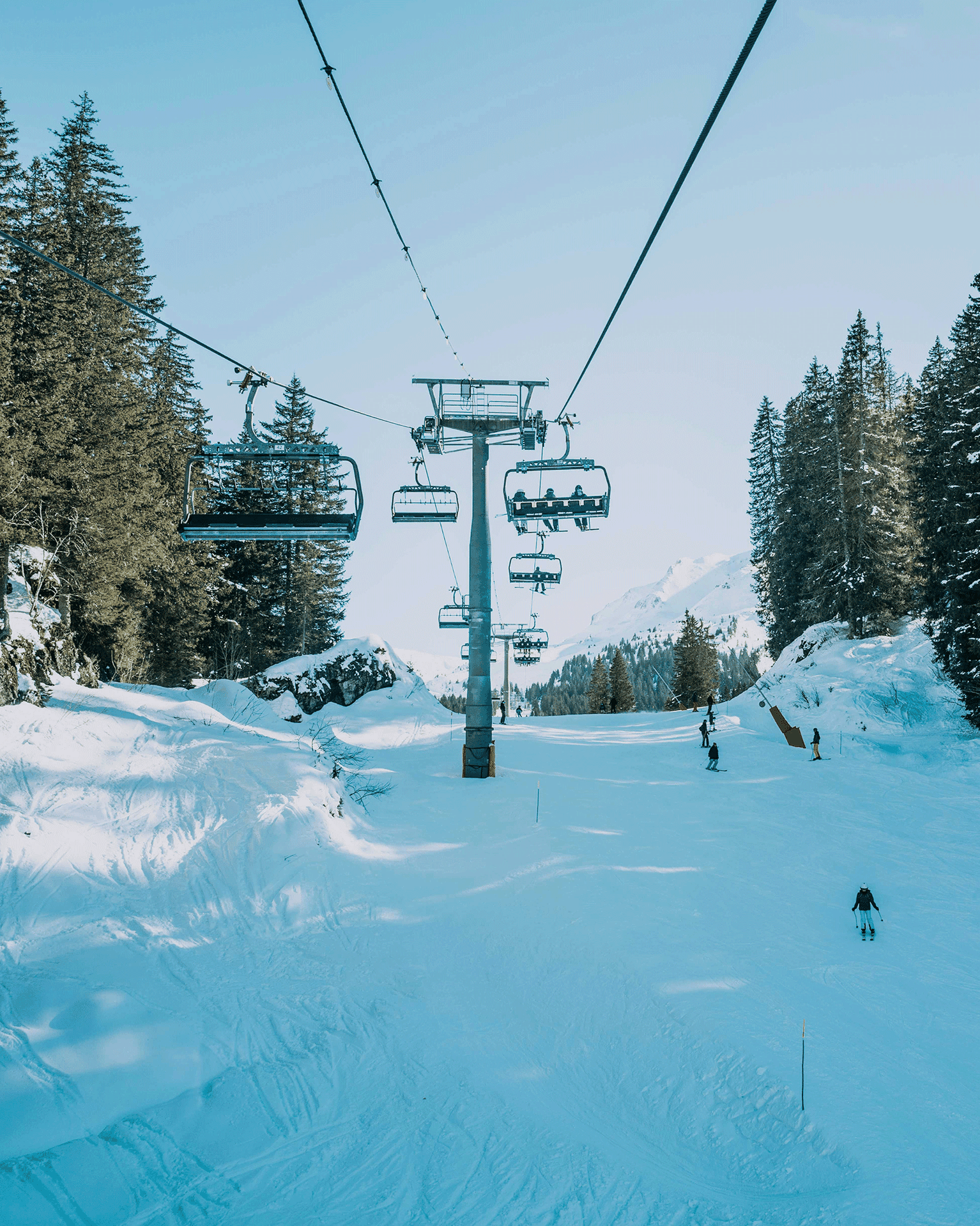Ski lift ascending a snowy mountain with skiers on a sunlit slope surrounded by pine trees.