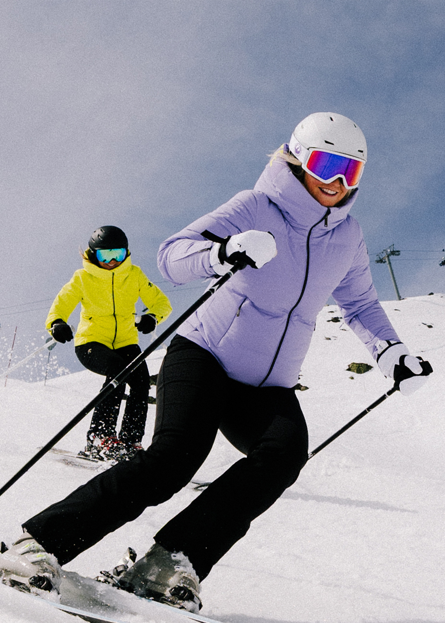 Two skilled skiers carving a snowy slope; lead in lilac jacket and white helmet, follower in bright yellow jacket and black trousers.