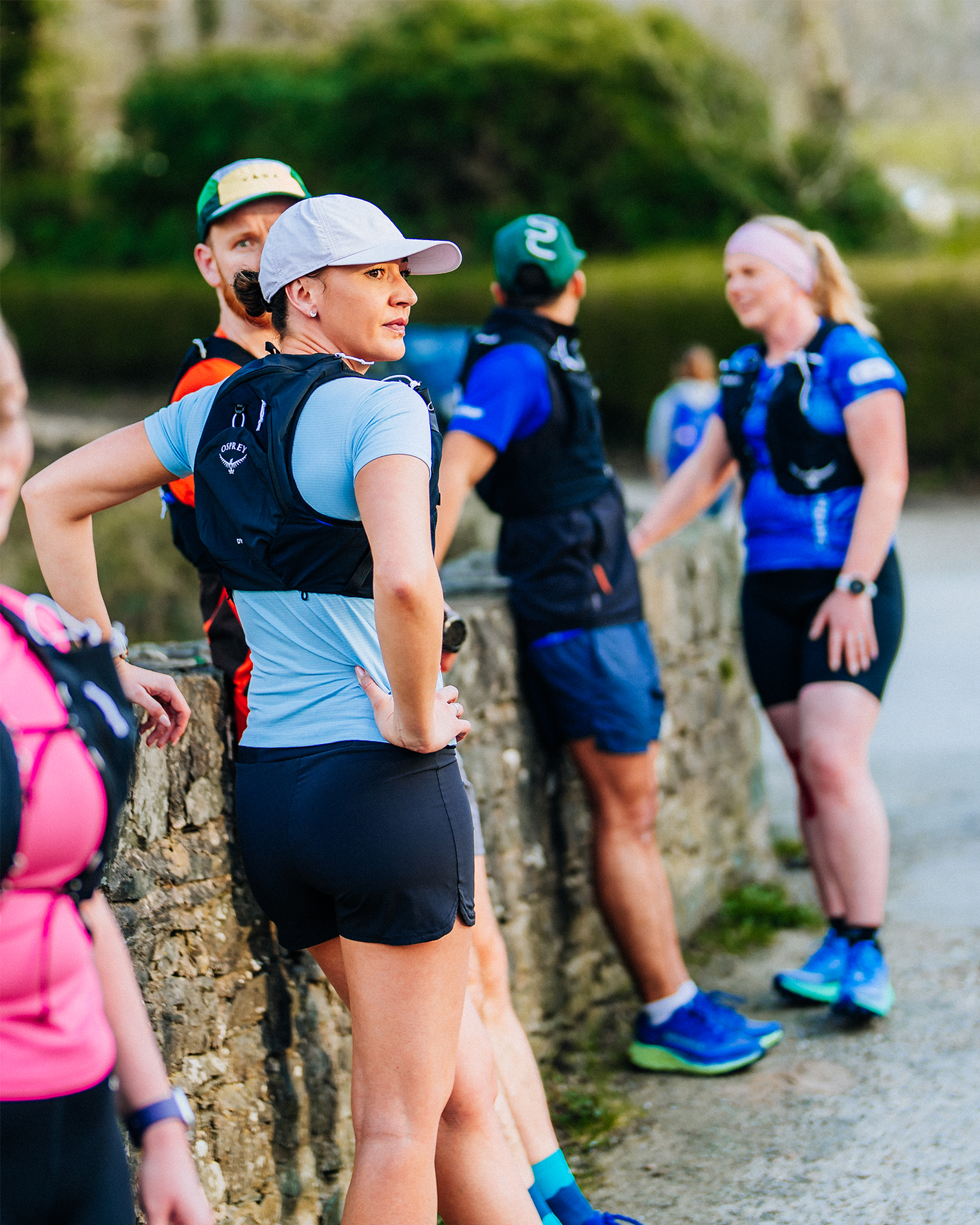 Group of runners wearing athletic gear and hydration packs, resting by a stone wall on a sunny day.