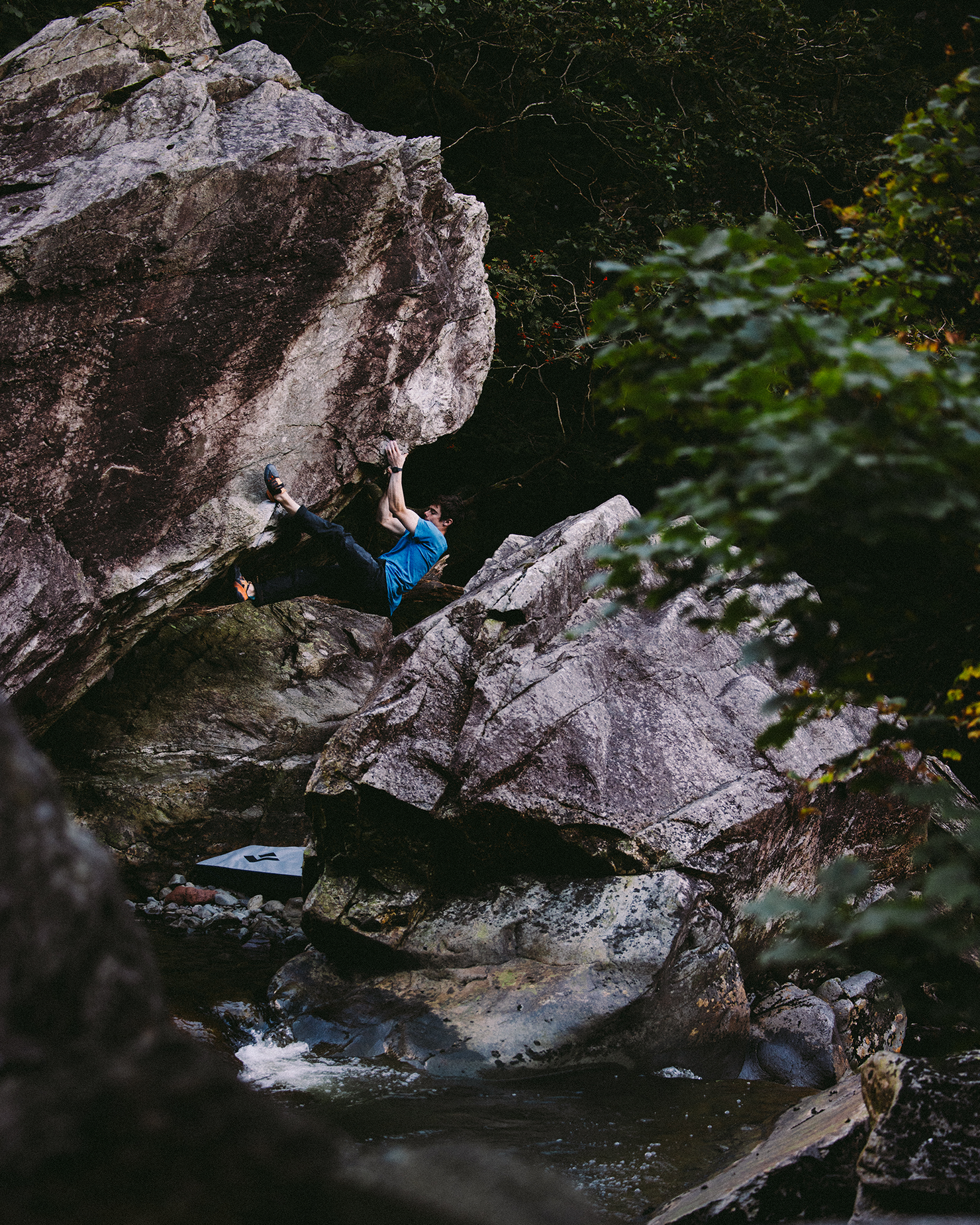 A climber bouldering on large rocks in a wooded area, wearing a blue shirt and climbing shoes, surrounded by trees and a small stream.