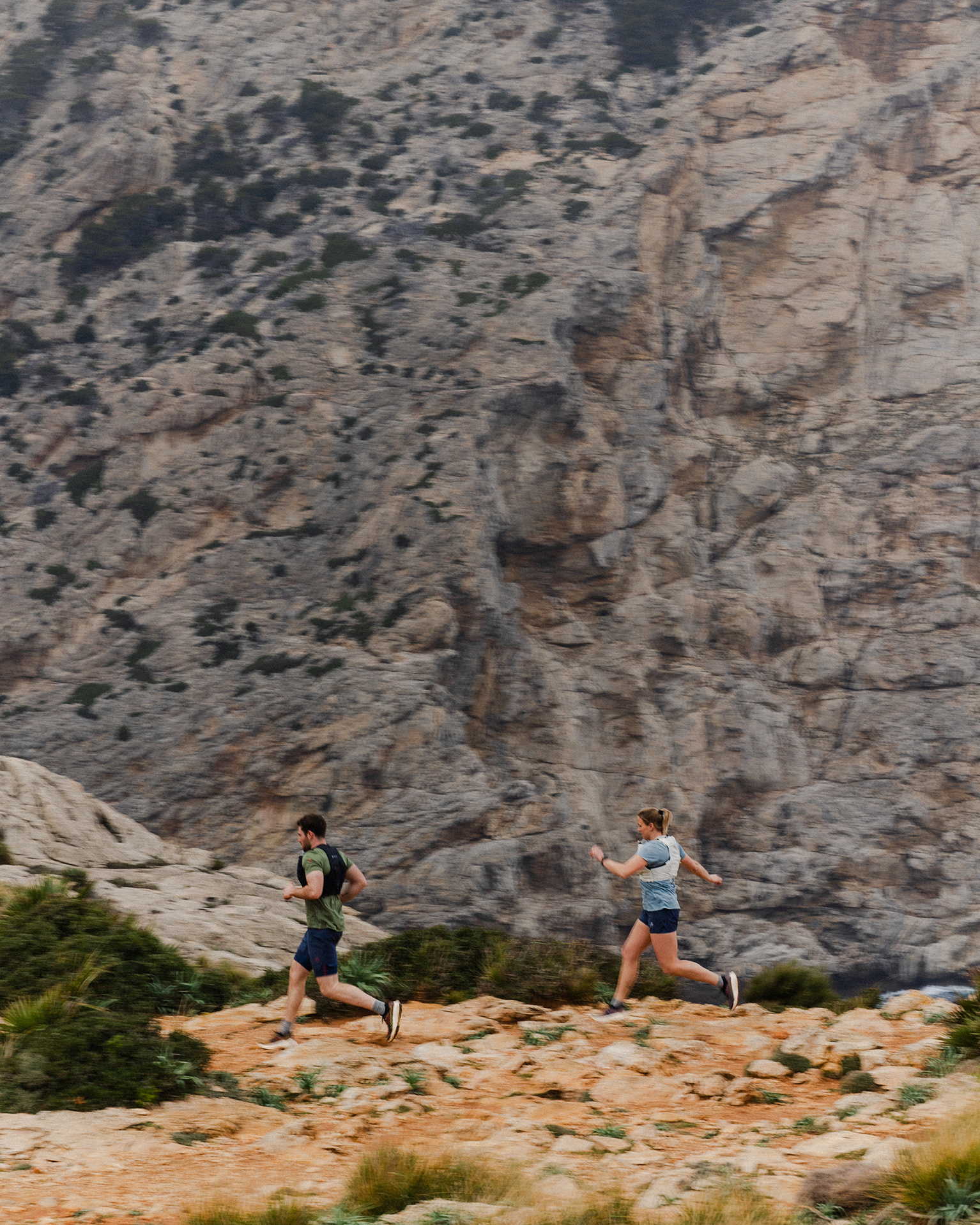 Two people trail running on a rocky mountain path with a rugged cliff in the background.