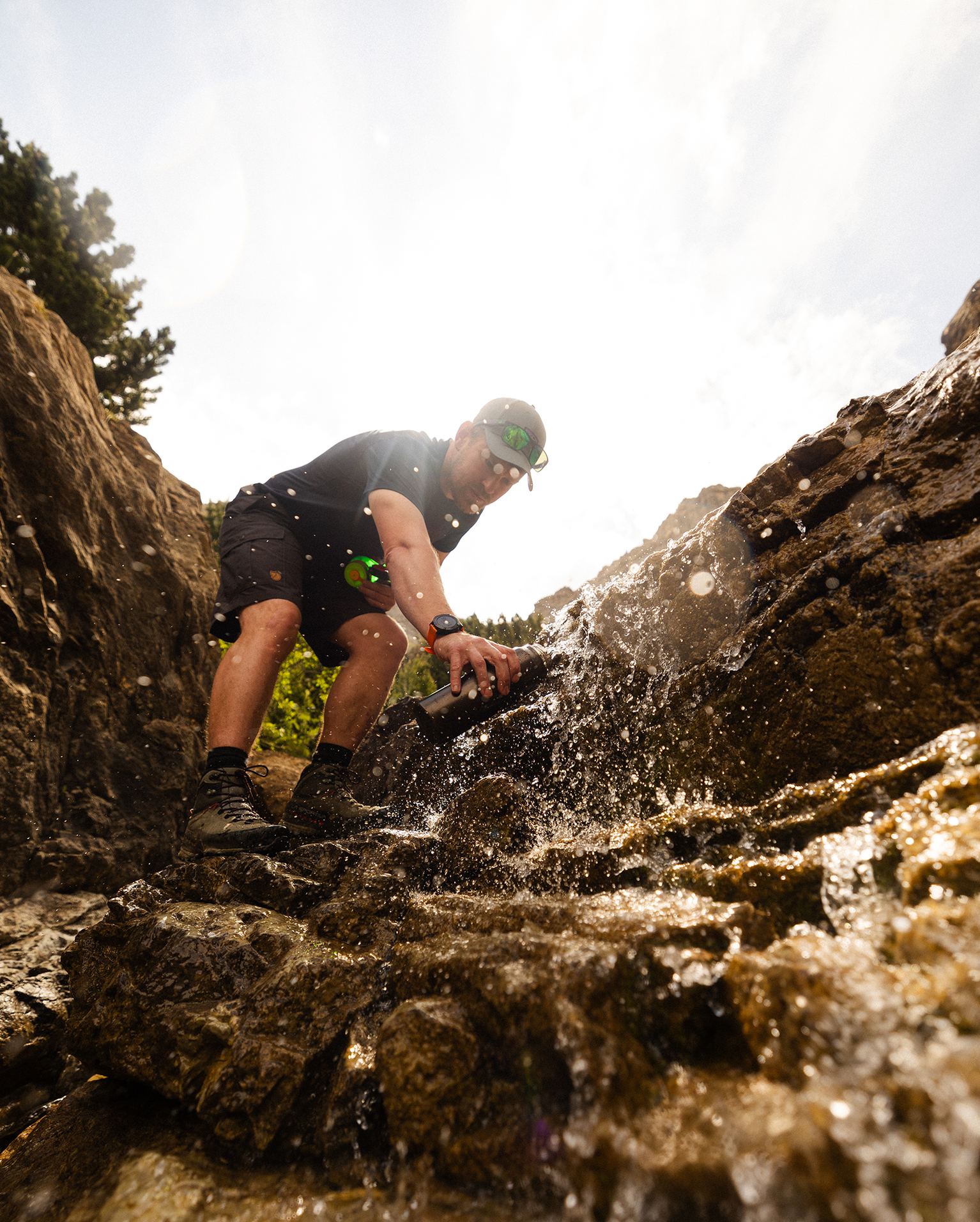 Man in outdoor gear leans over a rocky stream, splashing water. Sunlight shines through trees, creating a bright, adventurous scene.
