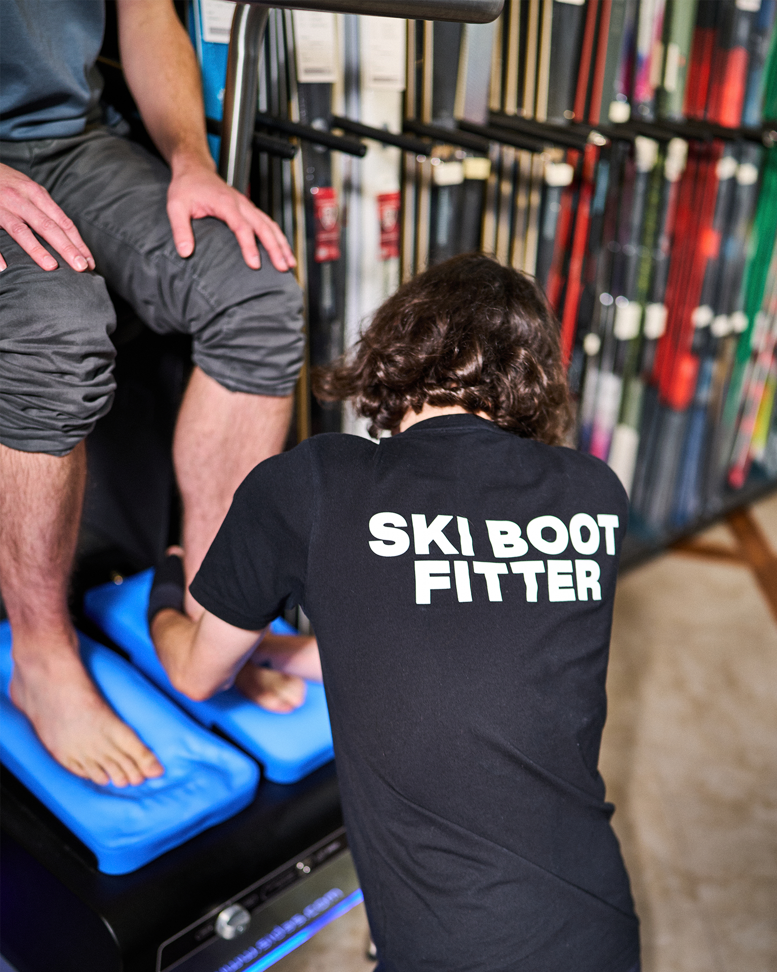 Person fitting ski boots for a seated customer, surrounded by ski equipment.
