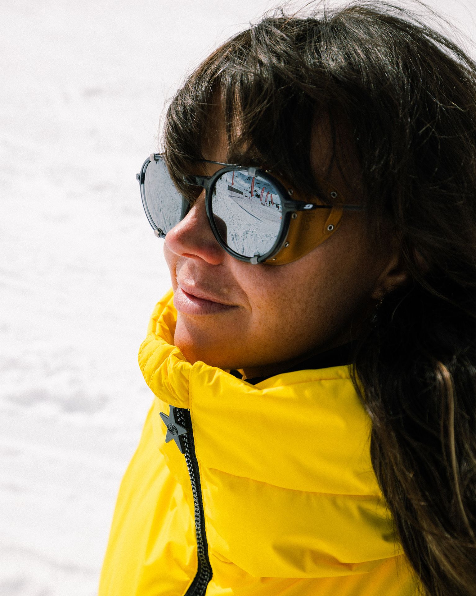 Woman wearing sunglasses and a bright yellow jacket, standing in a snowy landscape.