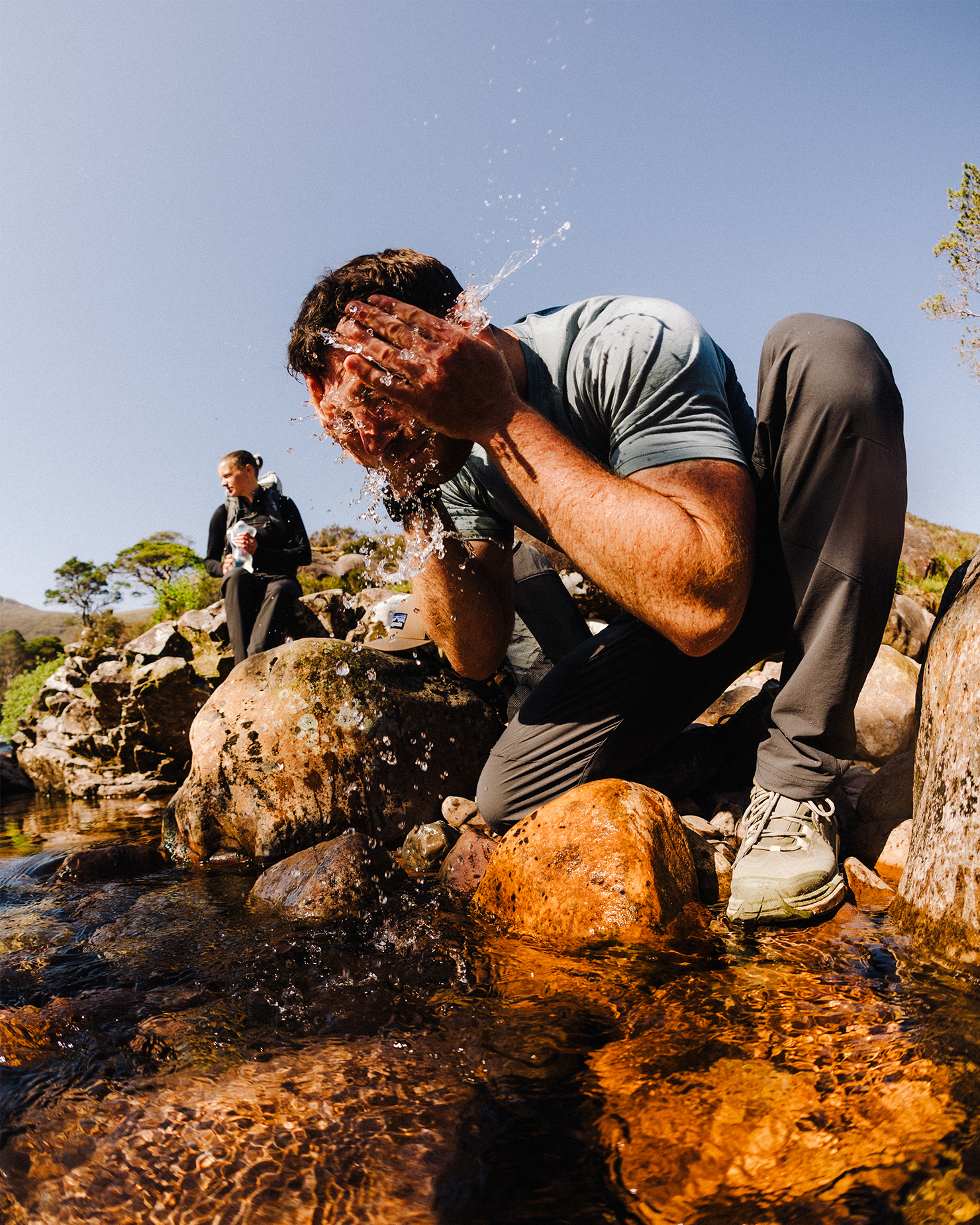 Man splashes water on face by a rocky stream, another person in background, clear sky above.