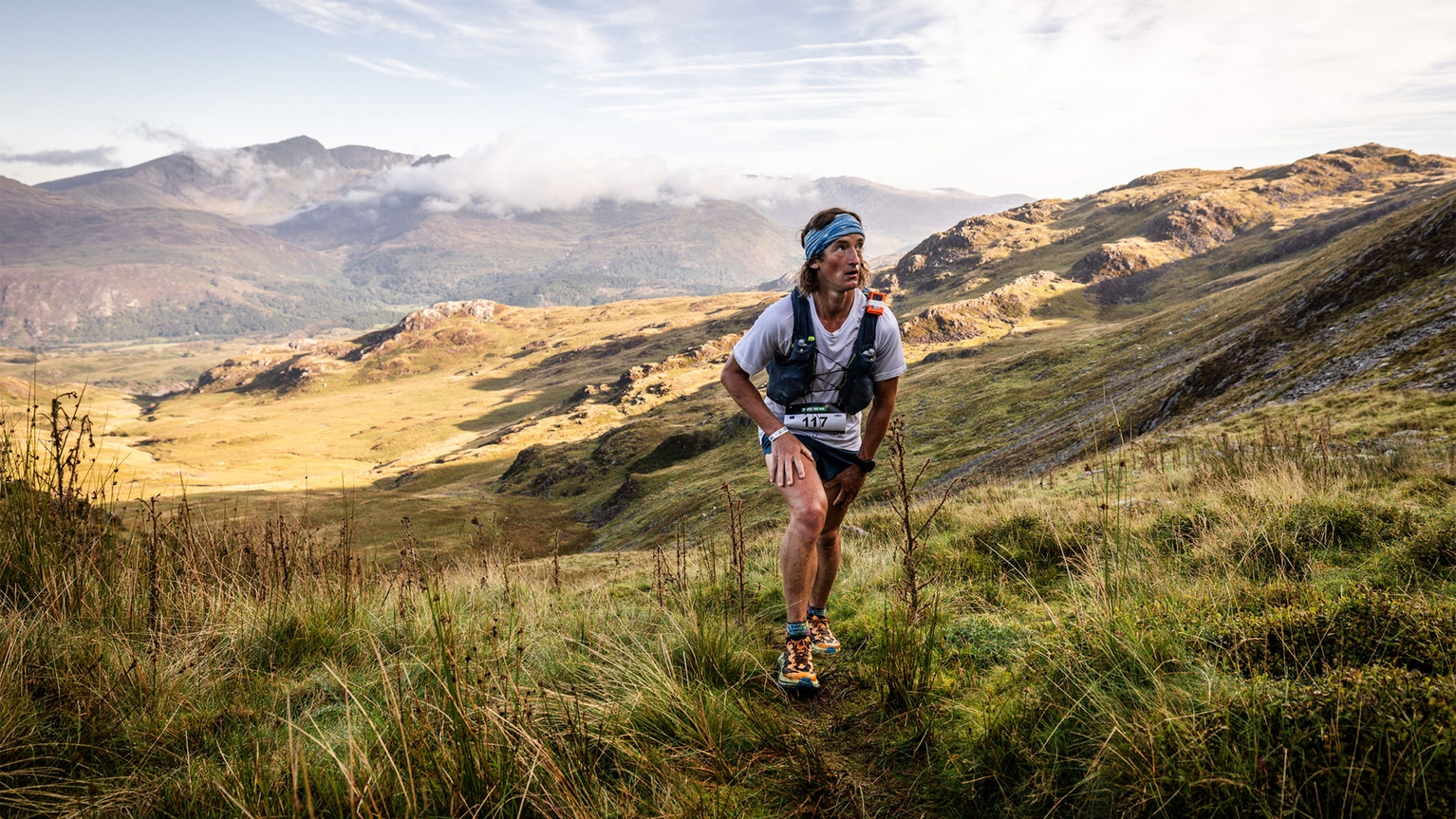 A runner in a race climbs a grassy hill with mountainous scenery in the background under a partly cloudy sky.