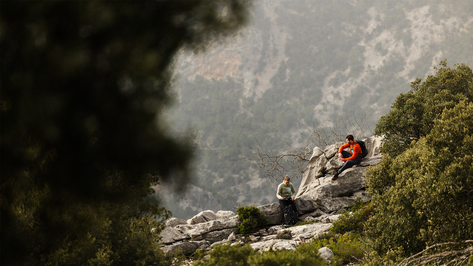 Two people in mountain gear rest on rocky terrain, surrounded by lush green foliage and a hazy, expansive mountain view.