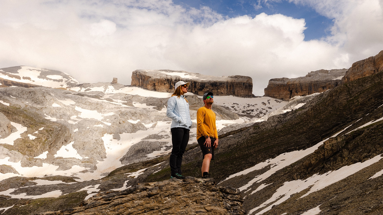 Two people stand on a rocky mountain ledge, surrounded by snow-dusted peaks and a cloudy sky.